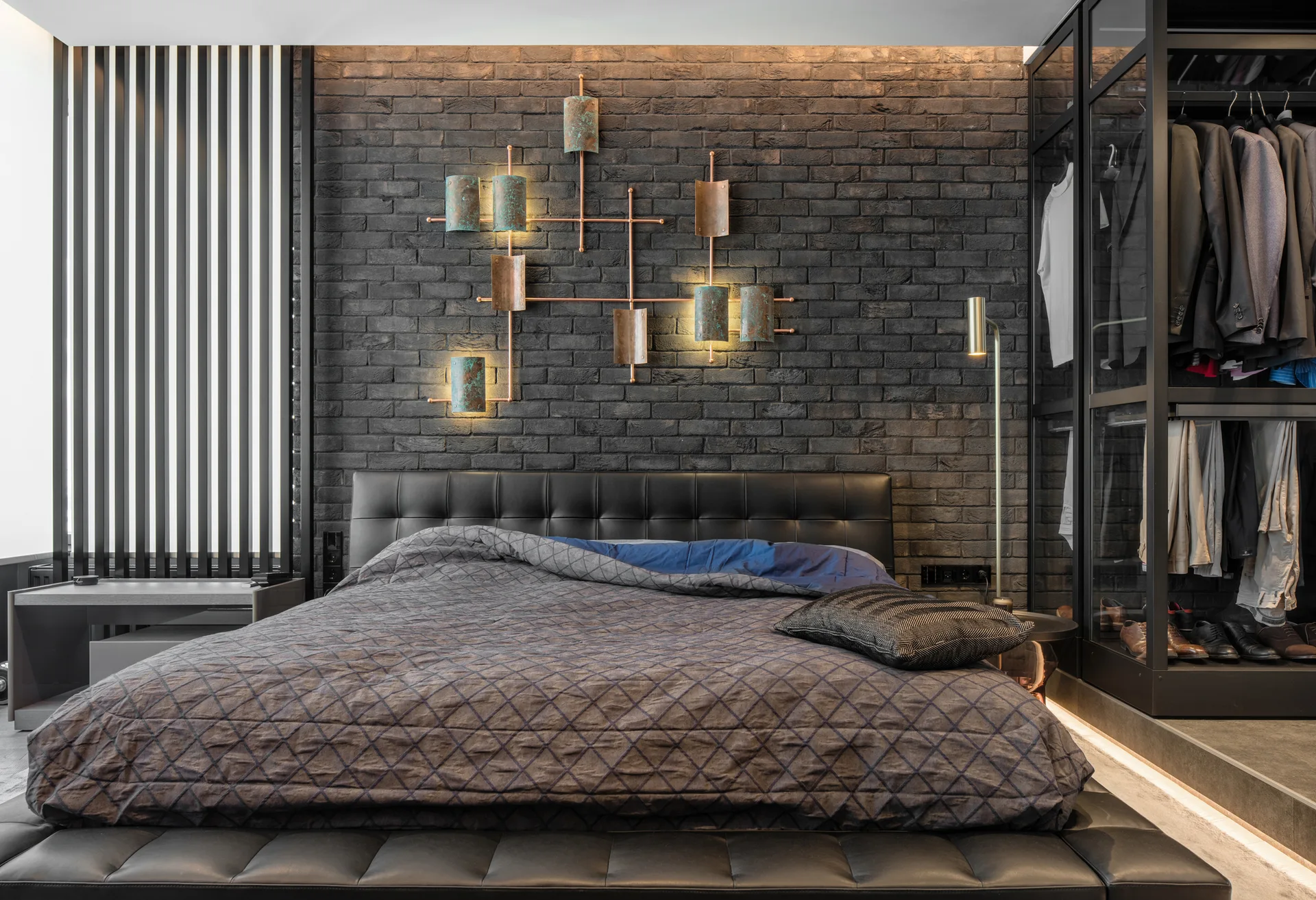 A dark modern bedroom featuring a charcoal brick wall with an illuminated copper sculptural fixture above a black tufted leather bed.