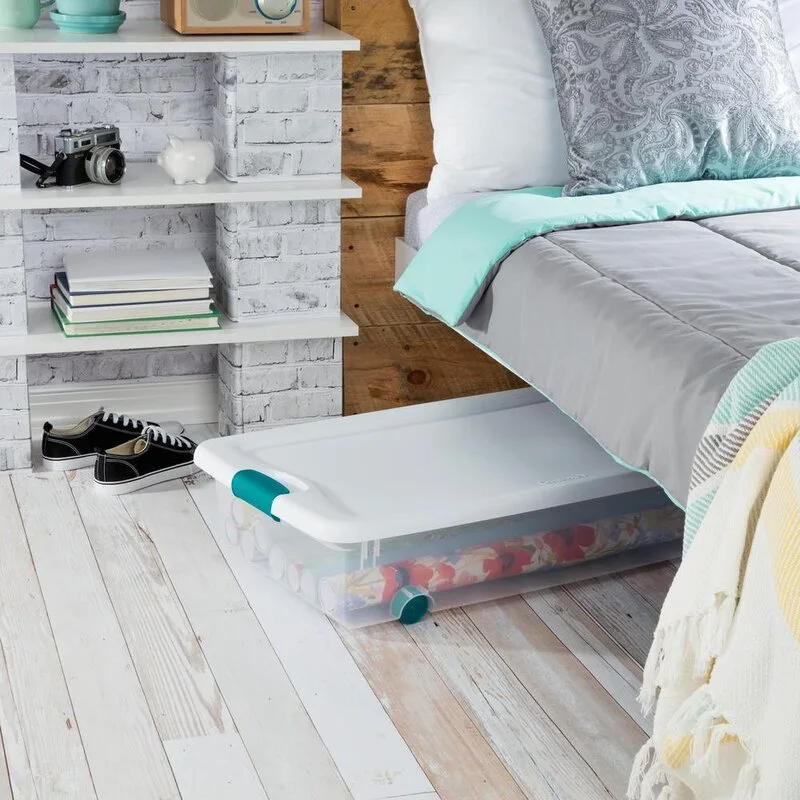 A clear plastic rolling storage container with teal latches sits on a whitewashed wood floor under a bed.