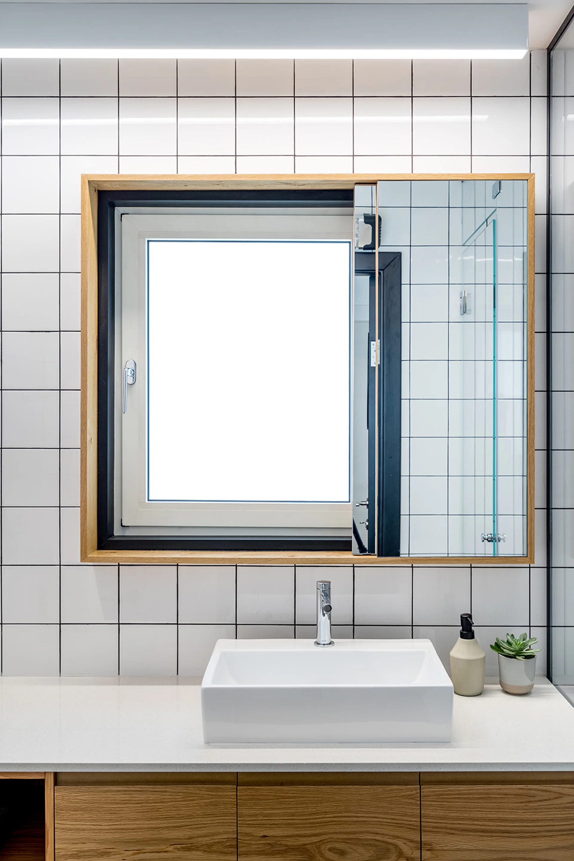 A modern bathroom vanity featuring a white square sink on a light wood cabinet, a white tiled wall, and a wood-framed window with a sliding mirror.
