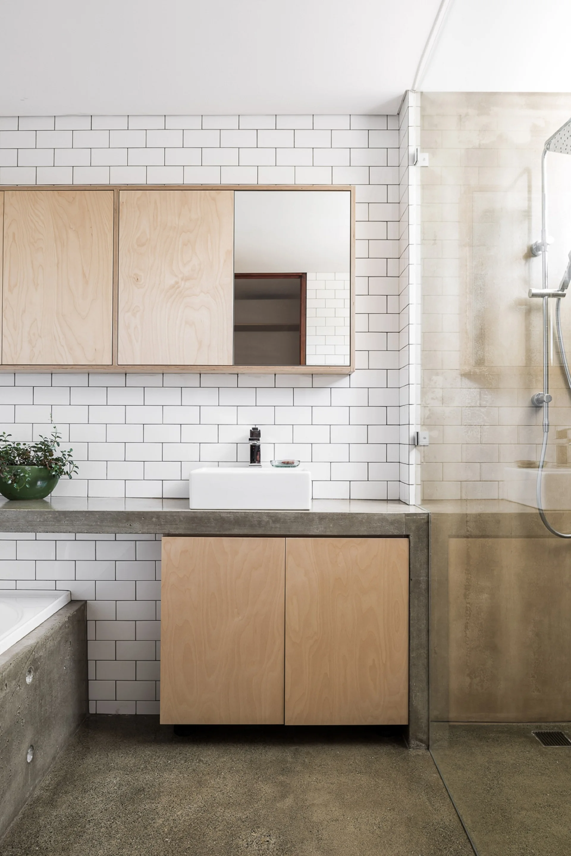 A modern bathroom featuring a concrete countertop with a white vessel sink, light plywood cabinets, and a white subway tile wall.