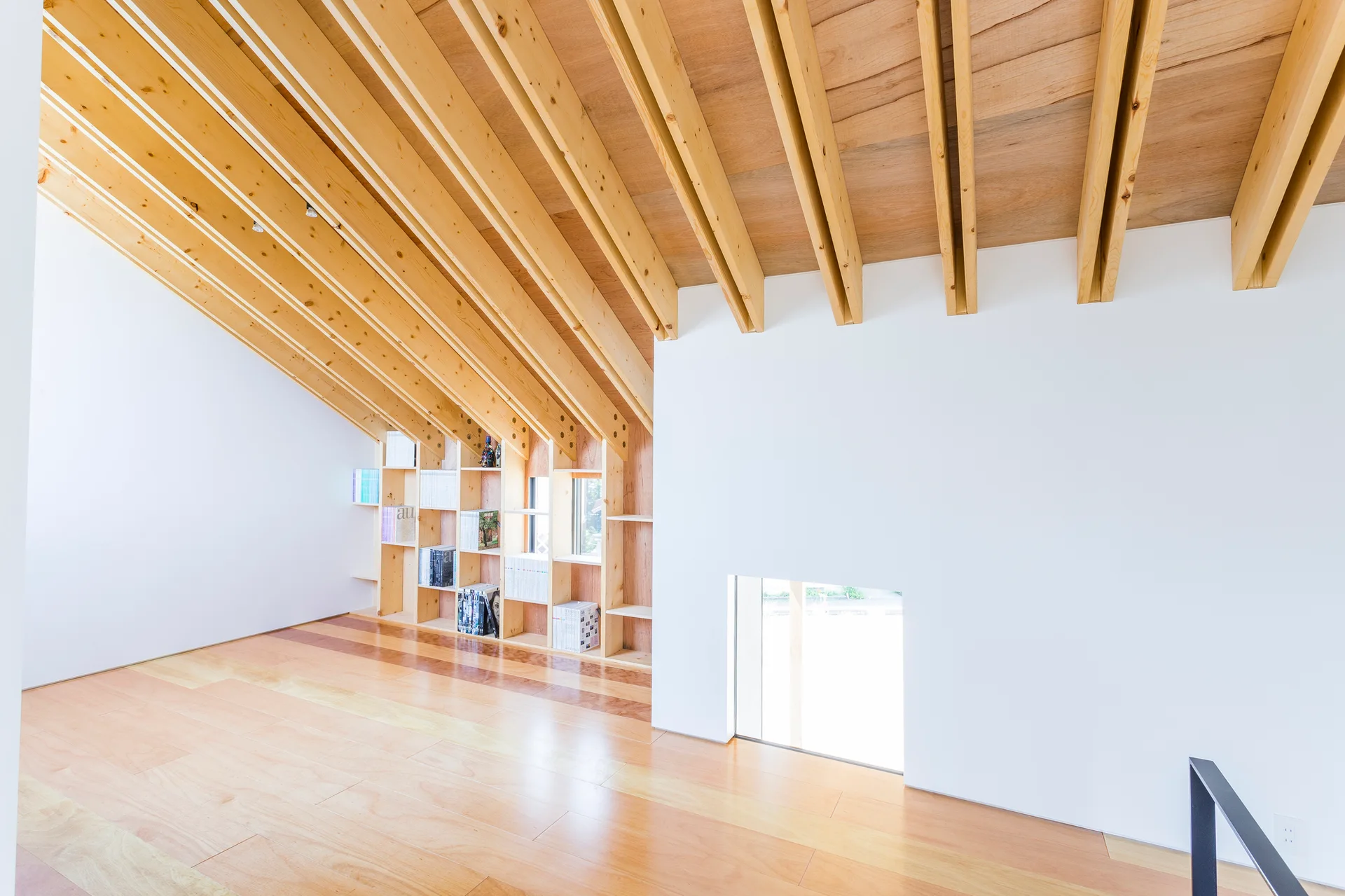 A minimalist attic space with a steeply sloped ceiling of exposed wood rafters, white walls, and a built-in wooden shelving unit.