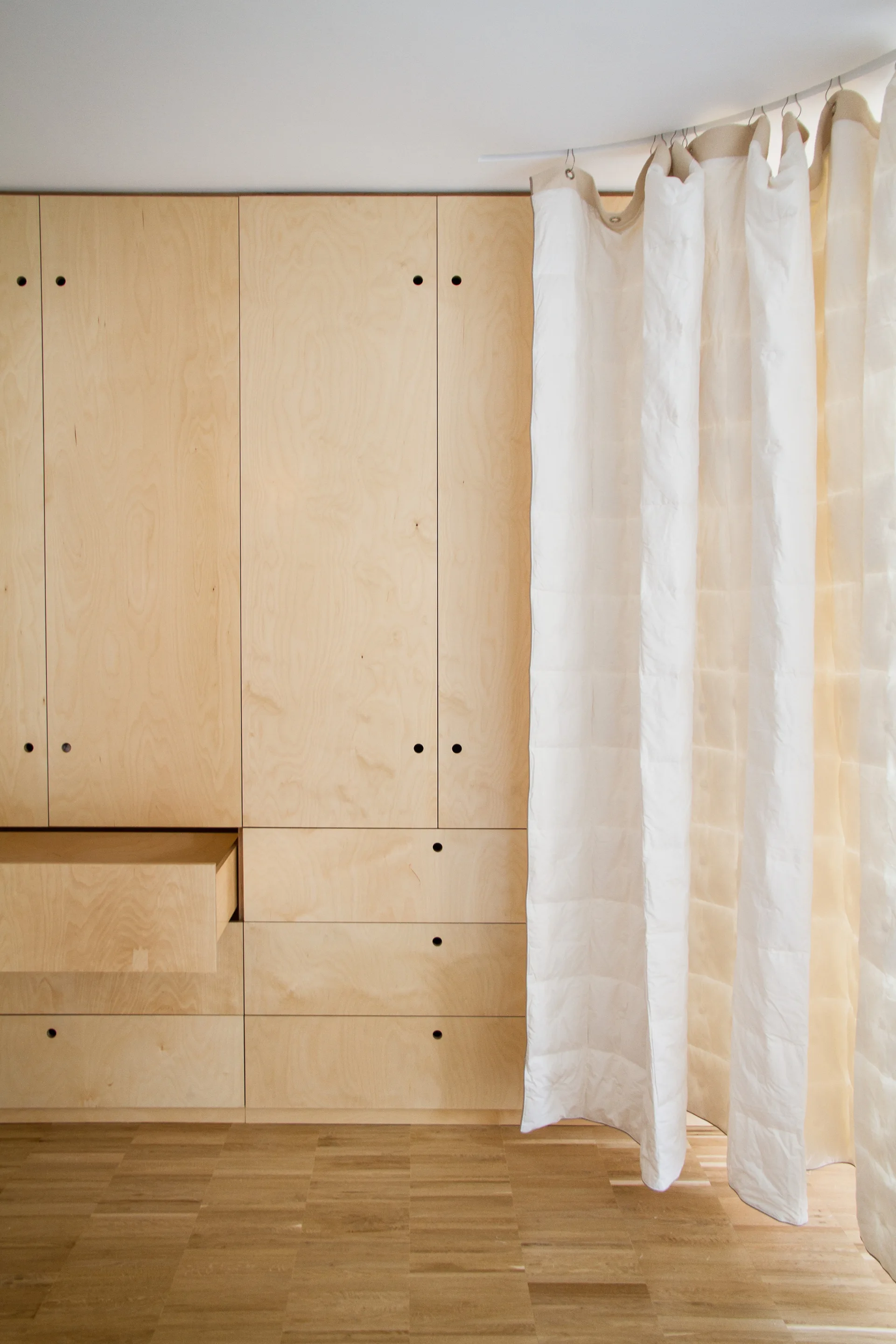 A wall of built-in plywood cabinetry with circular cutout pulls stands next to a white curtain hanging from a curved ceiling track.
