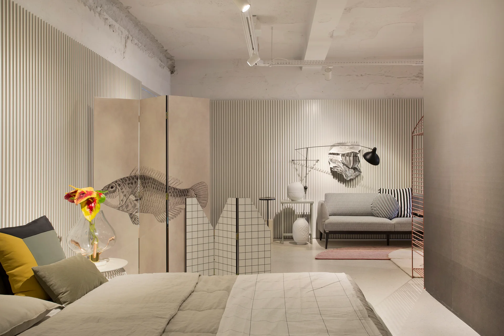 A contemporary studio bedroom features a bed, vertical ribbed wall paneling, a gray sofa, and illustrated folding screens used as room dividers.