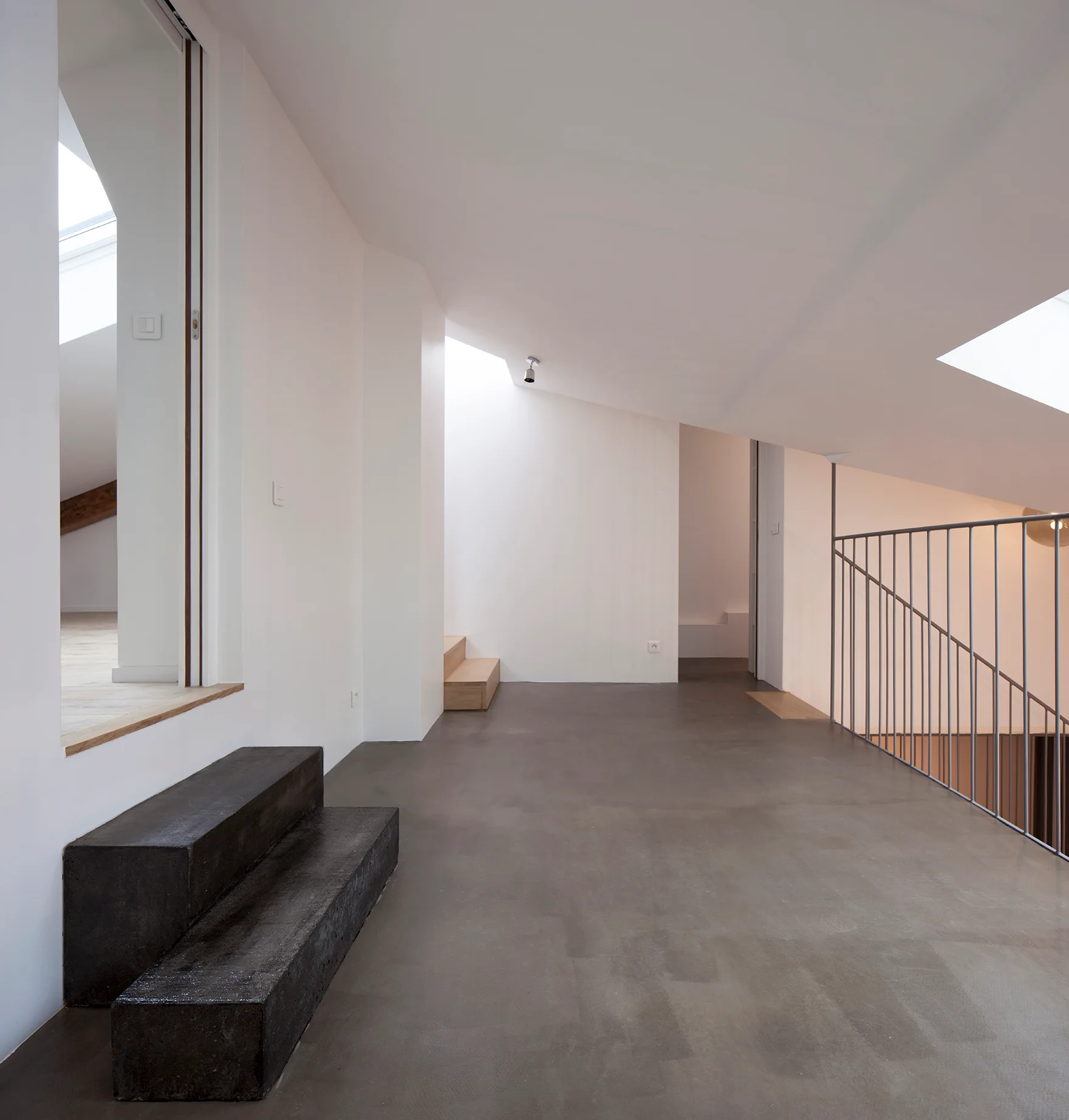 A minimalist attic landing features grey concrete flooring, white walls, a sloped ceiling with a skylight, a thin metal railing, and dark block steps.