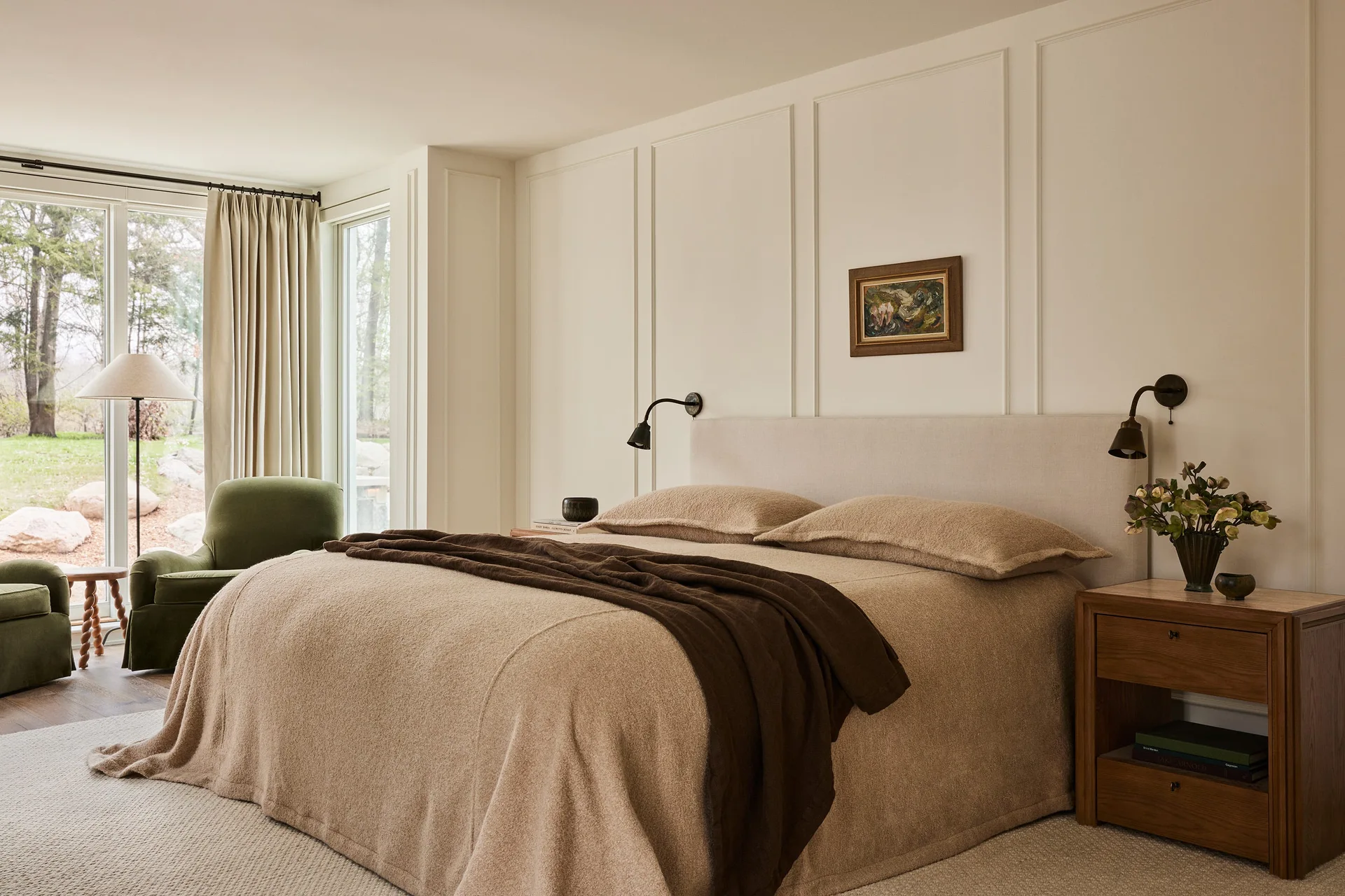 A bedroom features a bed with beige textured bedding and headboard against cream paneled walls, flanked by wooden nightstands.