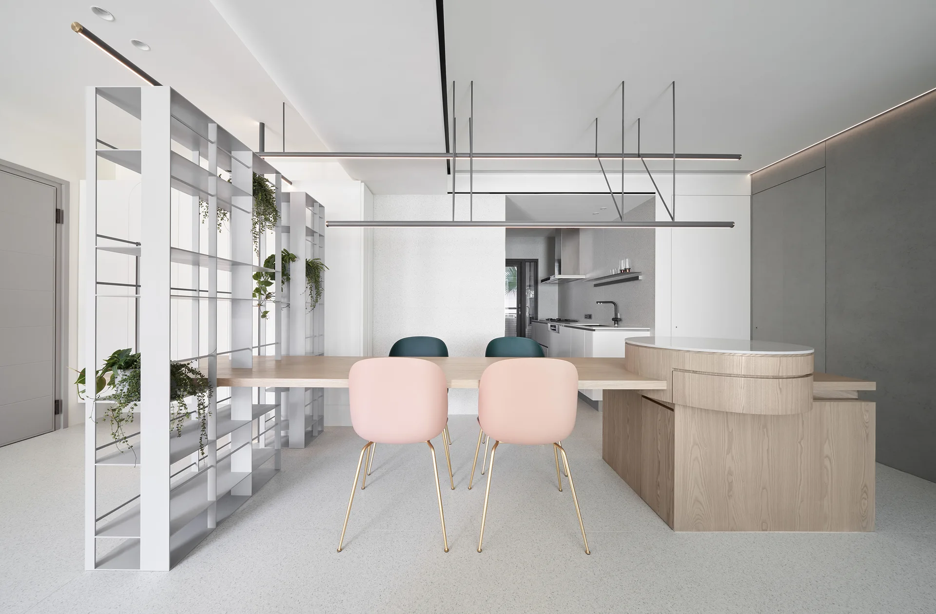 A modern dining room features a light wood table connected to a curved kitchen island, soft pink and green chairs, and an open metal shelving unit.