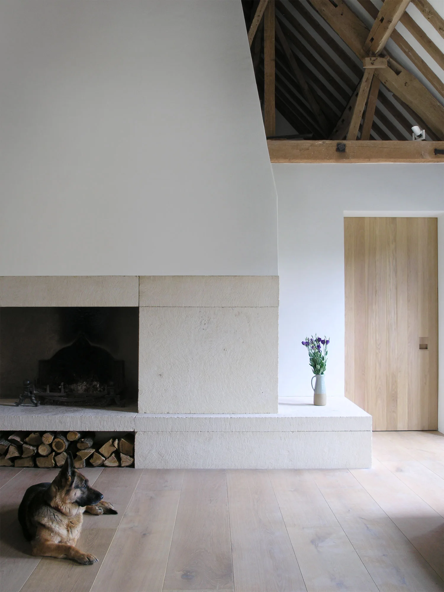 A large stone fireplace with integrated log storage stands on light wood floors near exposed wooden rafters and a flush door.