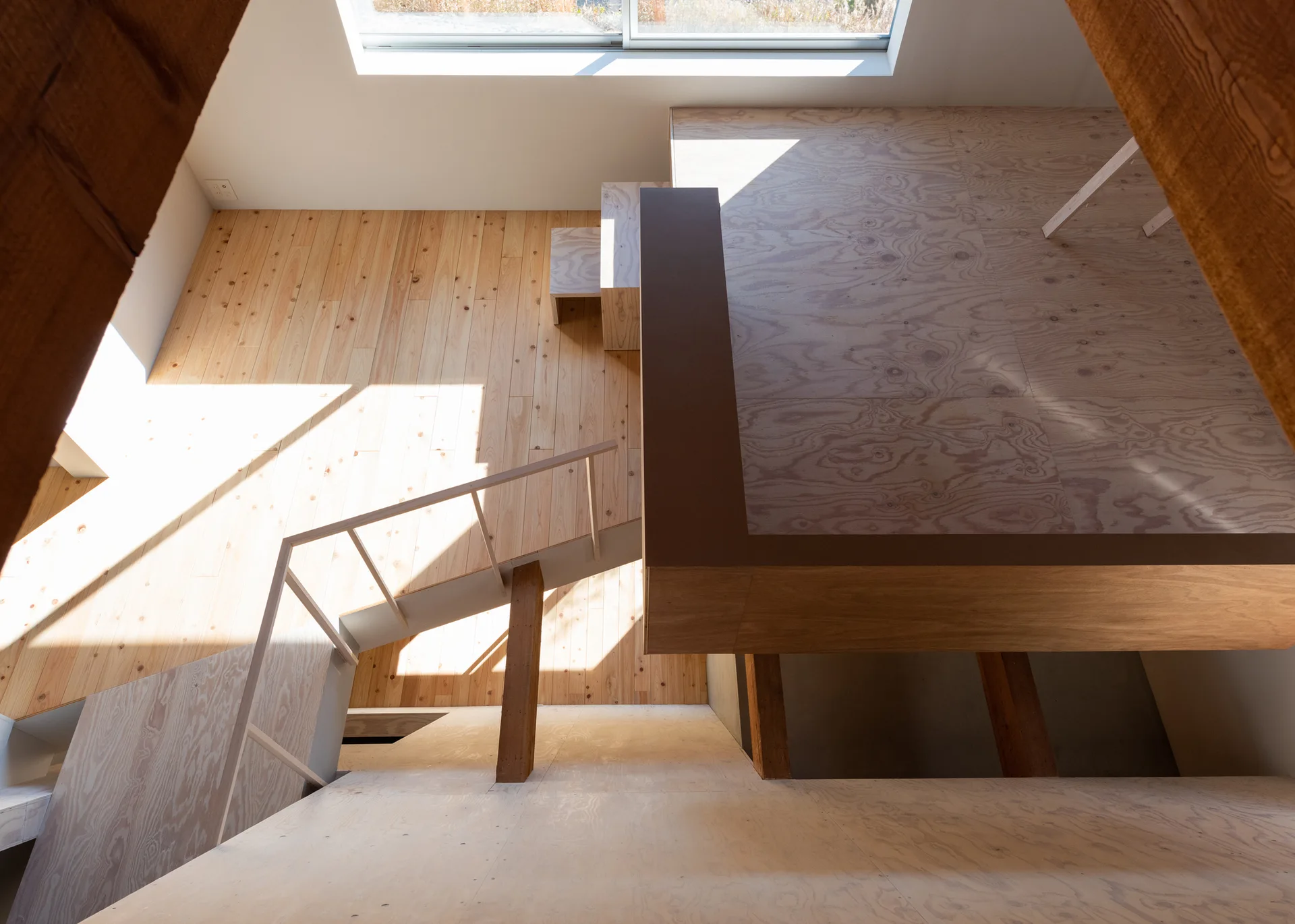 A high-angle view of a minimalist wooden house interior featuring a plywood mezzanine, a wood plank floor, and a central staircase with a slim railing.