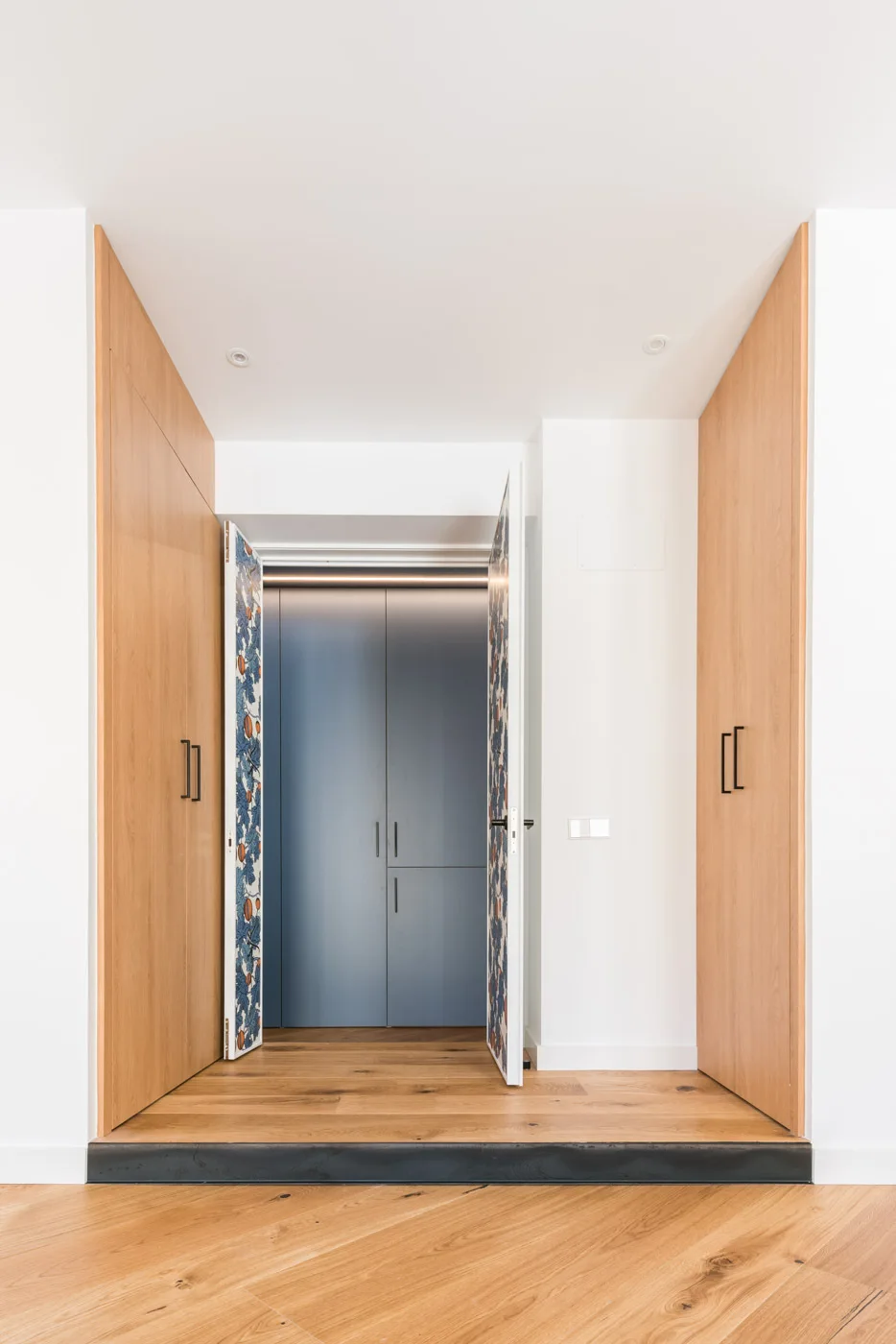 A minimalist entryway features floor-to-ceiling wood closets and an open doorway revealing blue cabinetry and doors lined with a blue botanical pattern.
