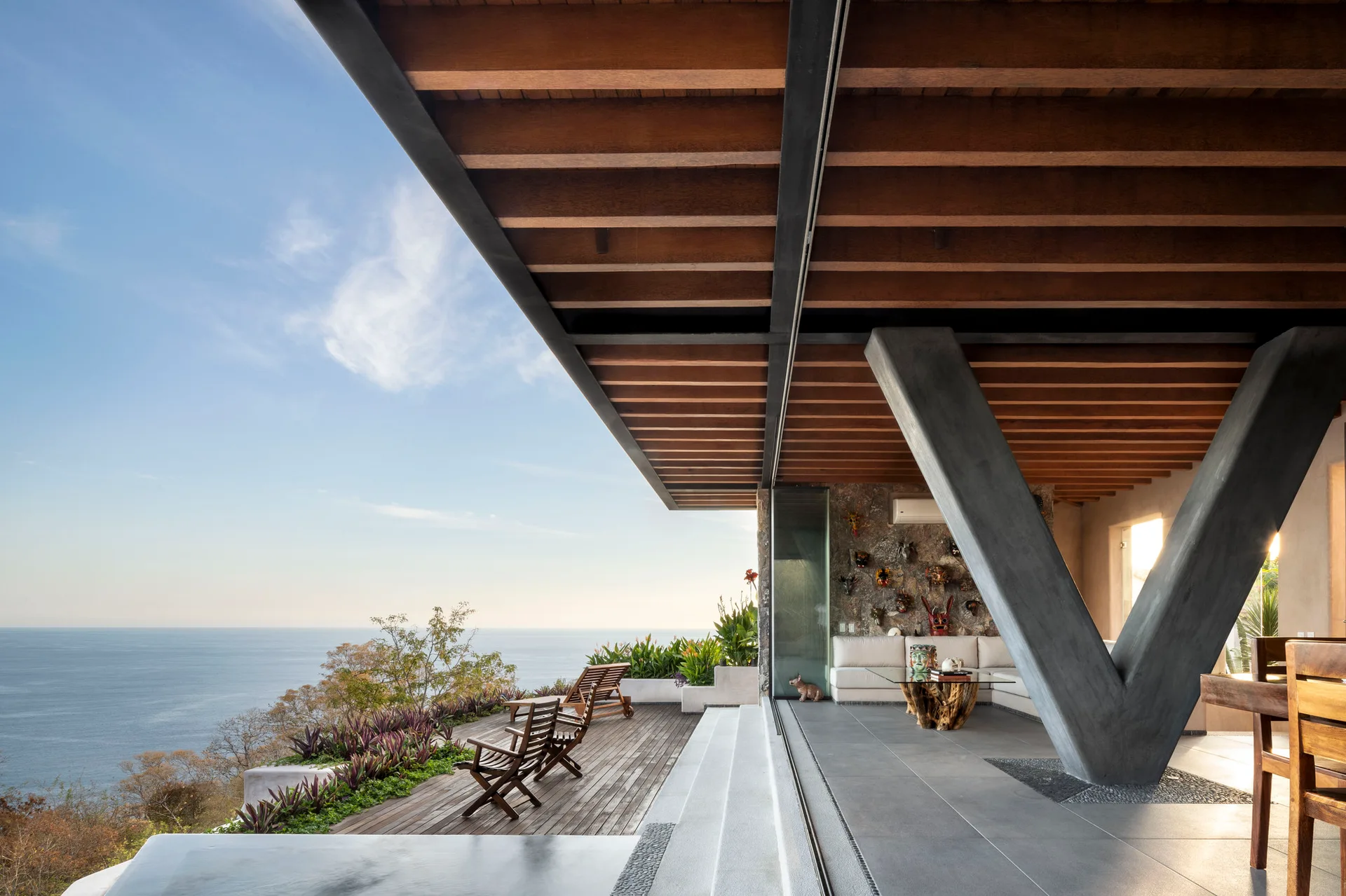 An indoor outdoor living room opens onto a wooden deck overlooking the ocean, featuring a massive V-shaped concrete pillar and exposed ceiling beams.