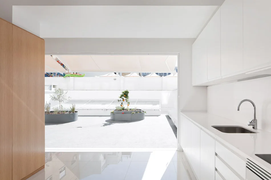A minimalist white kitchen with high-gloss cabinets and reflective tile flooring looking out onto a bright paved patio with circular planters.