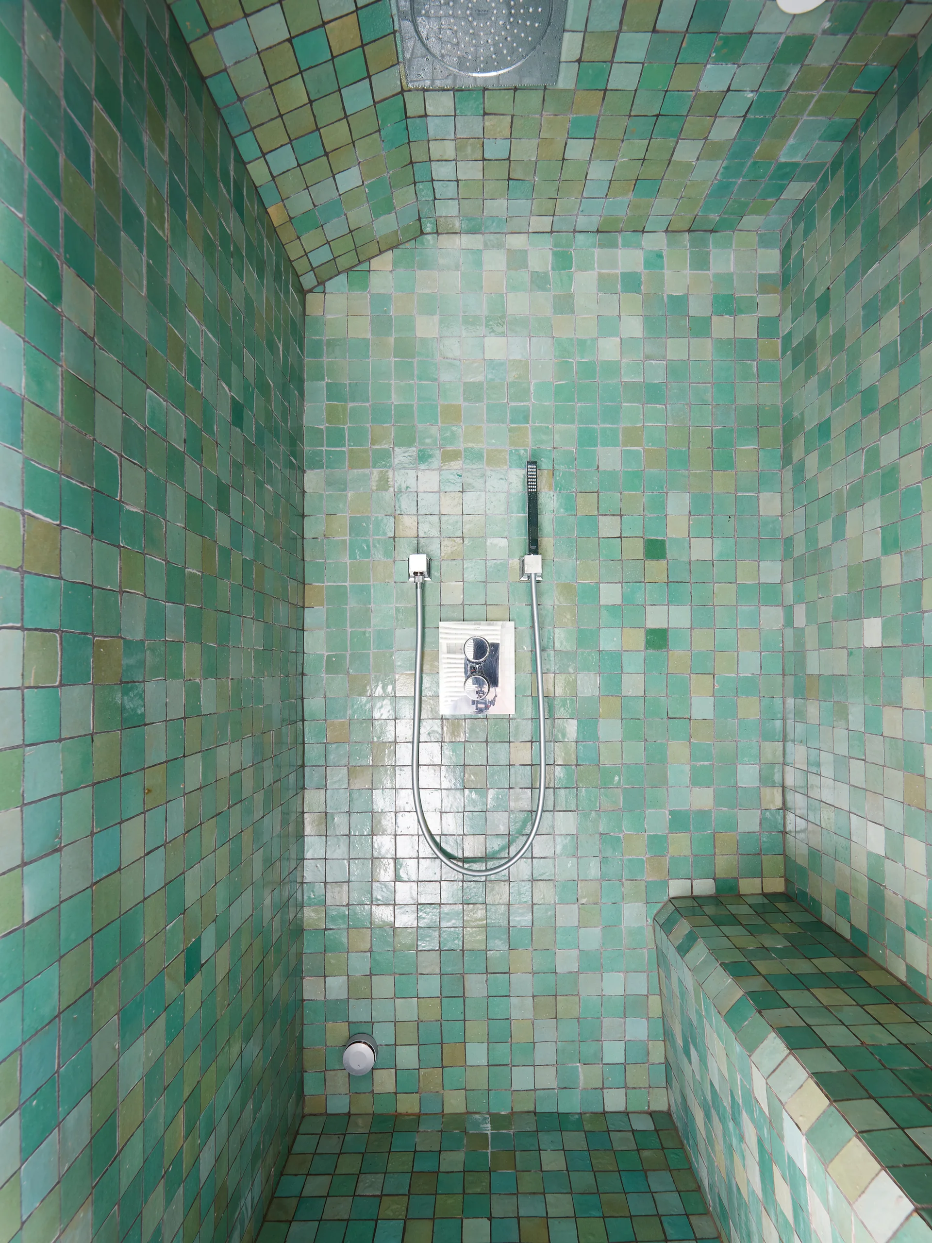 A walk-in shower completely covered in small, glossy green square tiles features chrome fixtures and a built-in tiled bench.