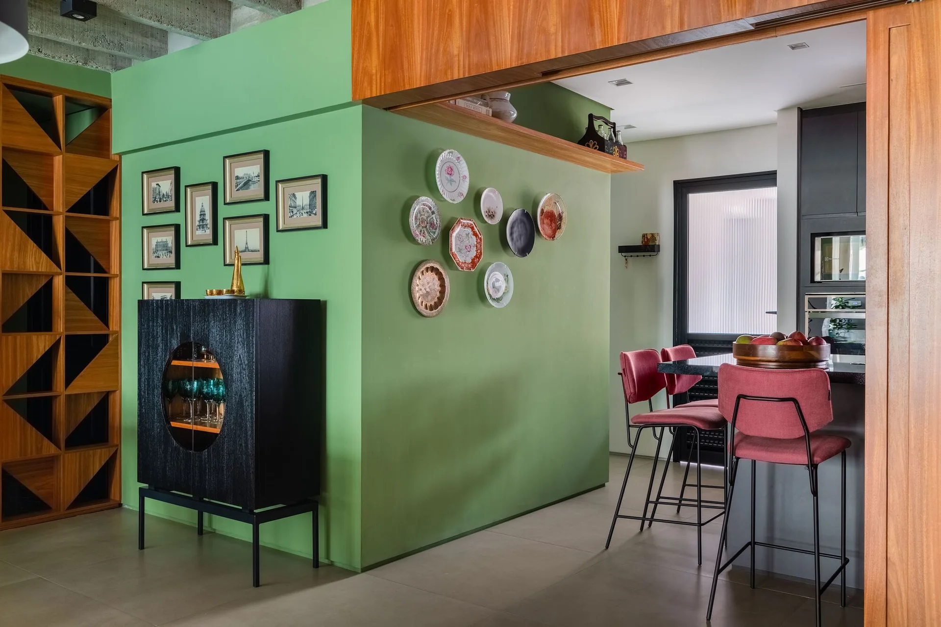 A green partition wall decorated with hanging plates separates a kitchen with red bar stools from an adjoining room with a black cabinet.