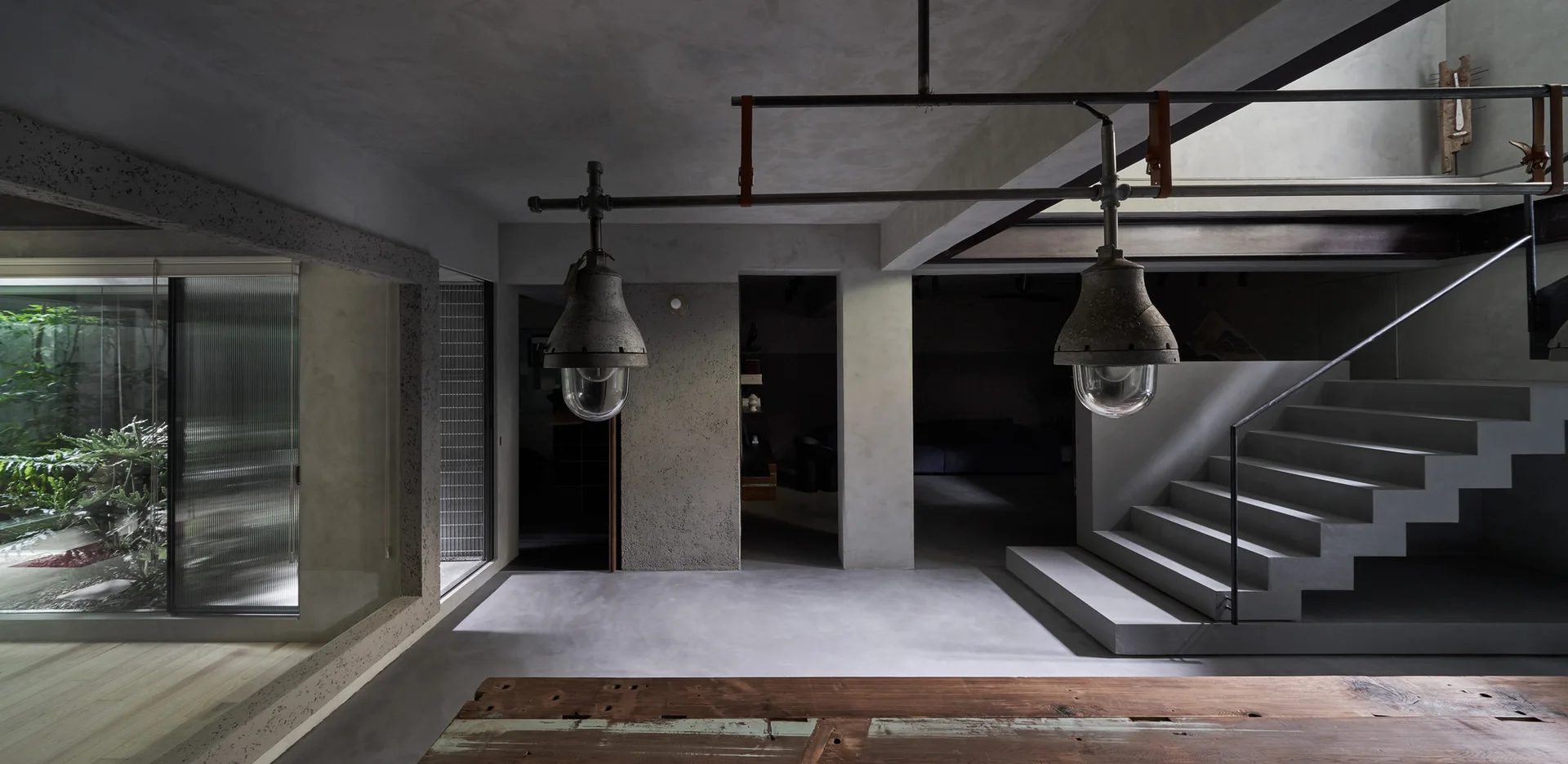 A minimalist concrete room featuring a floating staircase, heavy industrial pendant lights, and a rustic wooden table in the foreground.