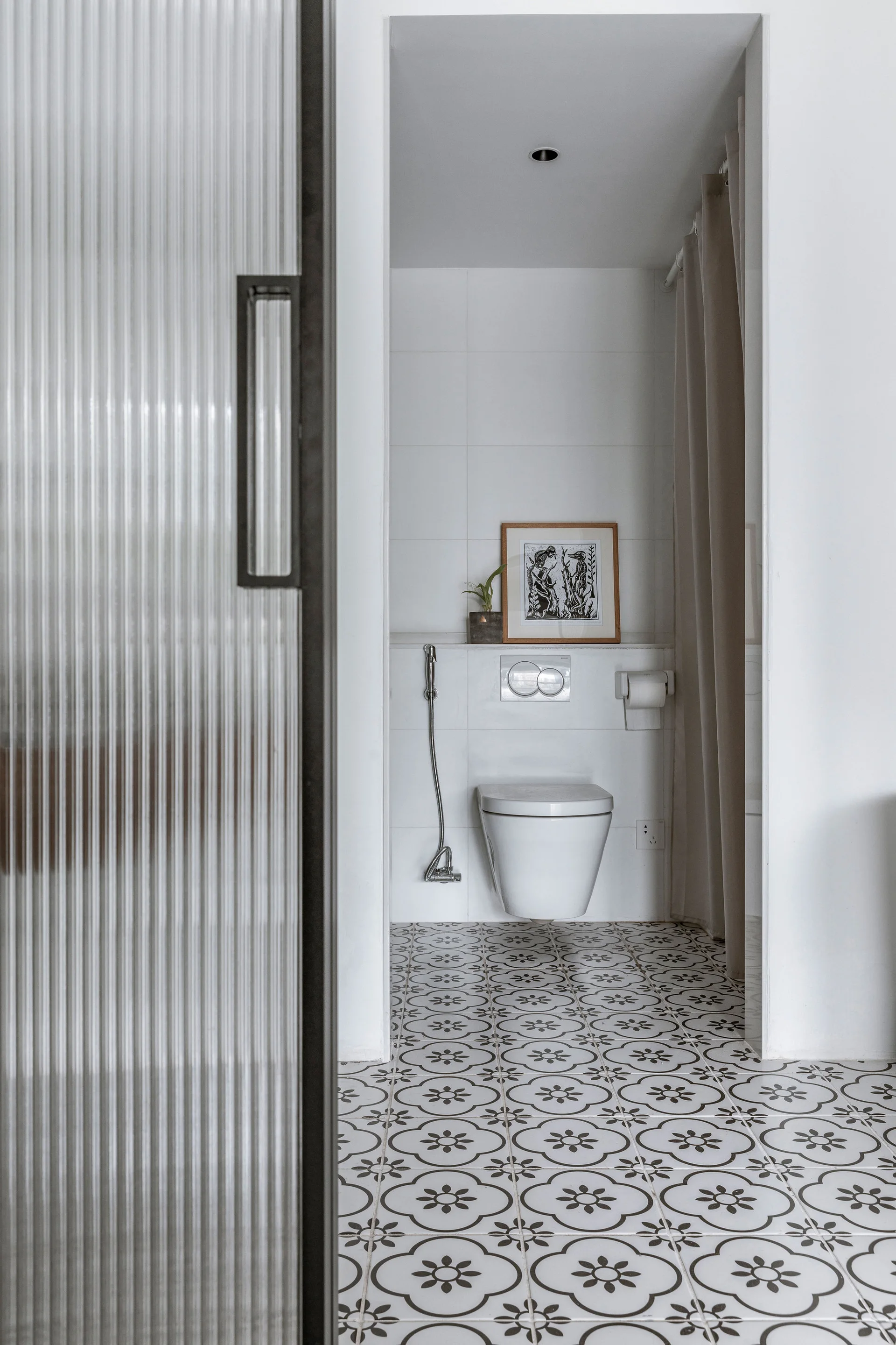 A modern bathroom features a wall-mounted toilet, black and white patterned floor tiles, and a partially visible fluted glass sliding door.