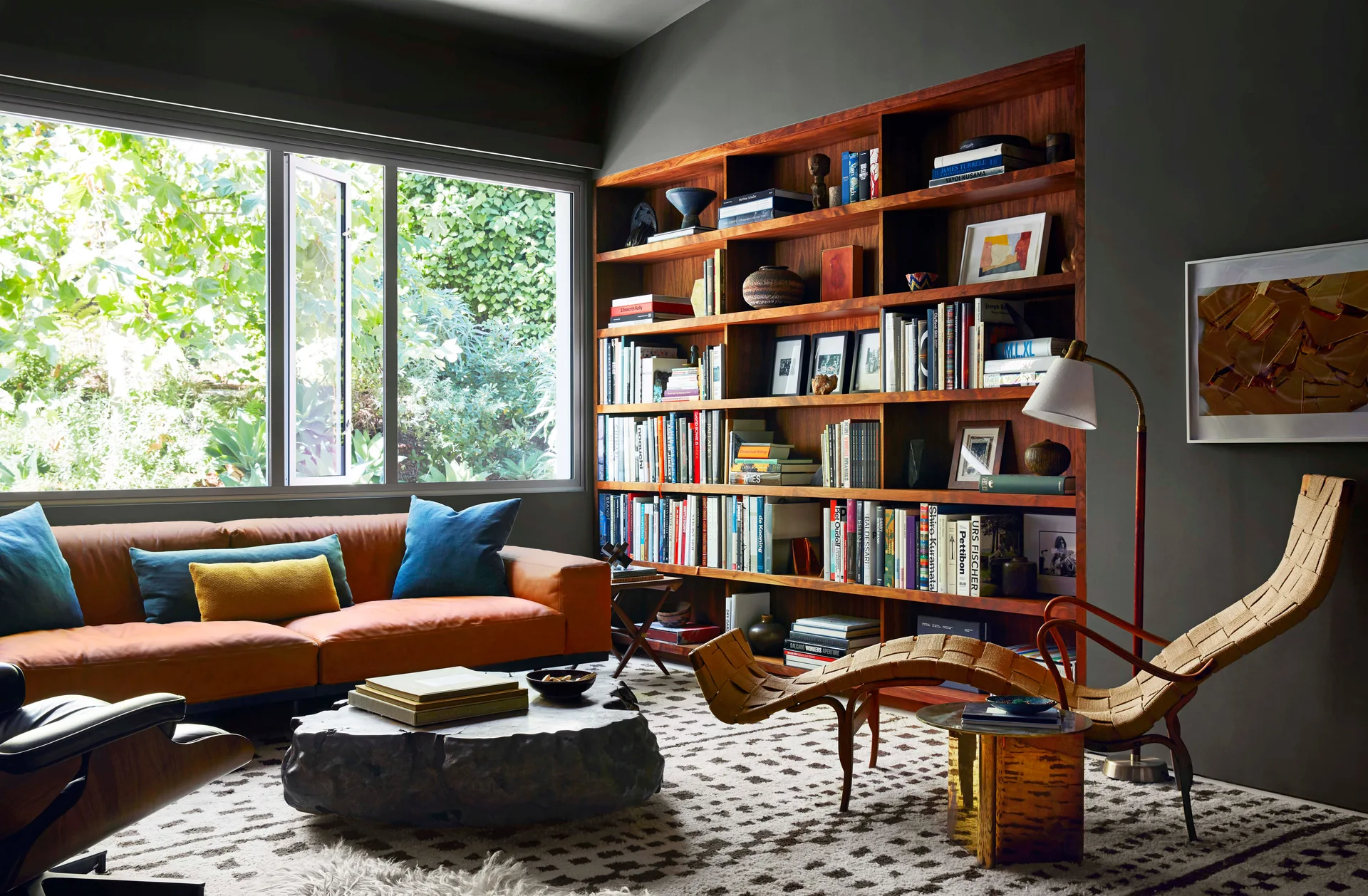 A living room with charcoal grey walls features a tan leather sofa, a built-in wooden bookcase filled with books, and a patterned geometric rug.
