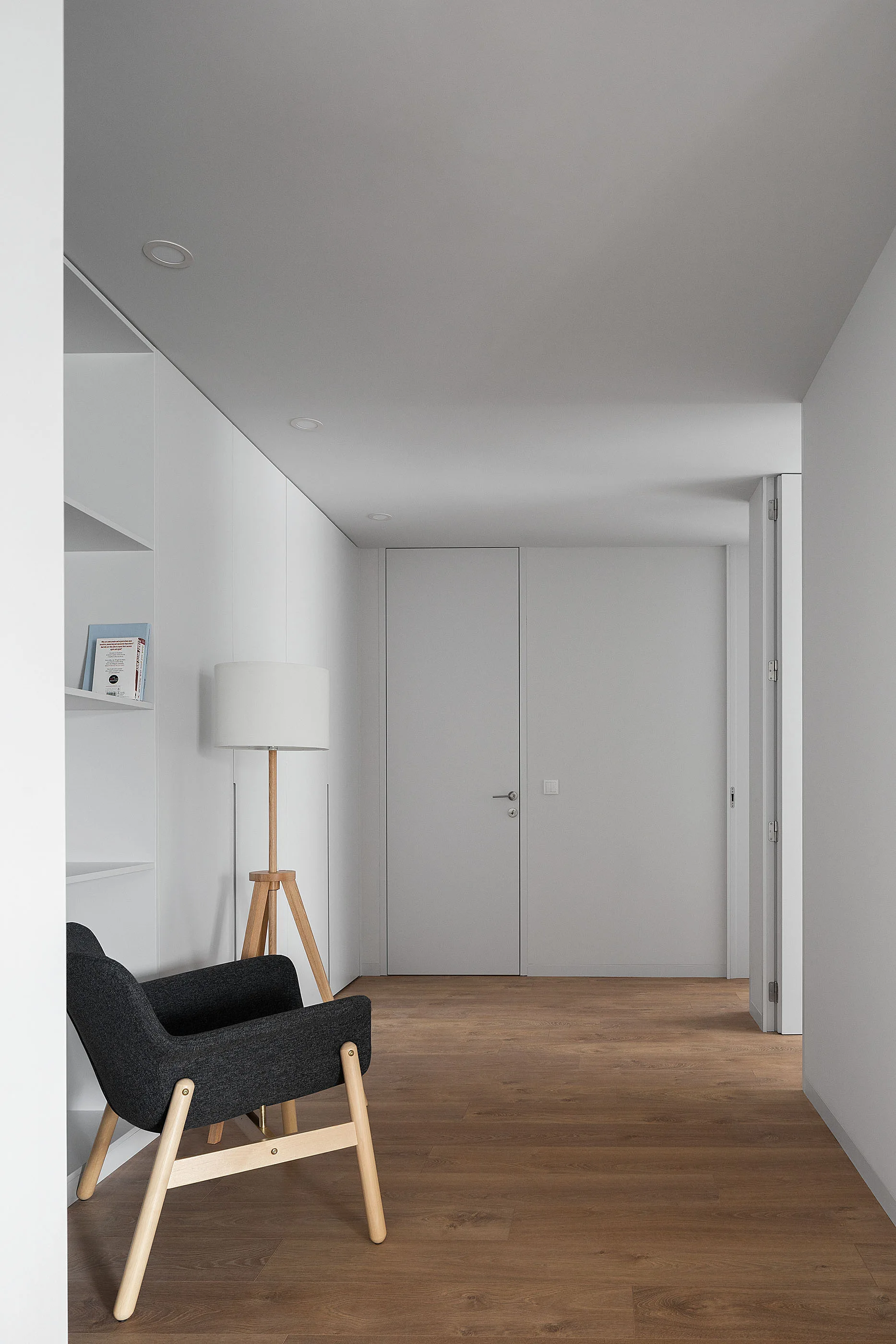 A minimalist white hallway features light wood flooring, a charcoal armchair, a wooden tripod floor lamp, and white built-in shelving.