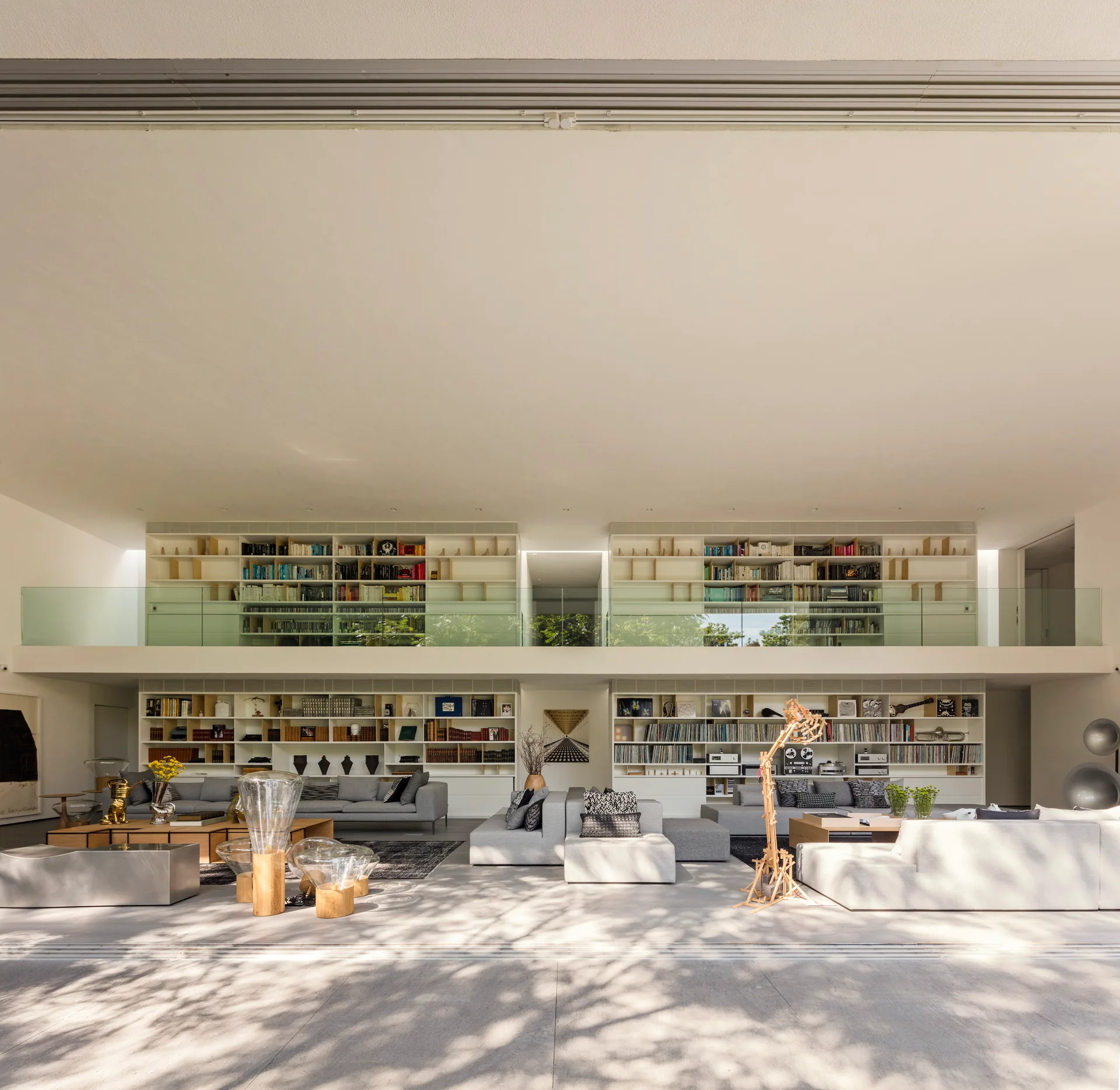 A double-height living room features white built-in bookcases, modular grey sectional sofas, and an upper mezzanine with a glass railing.