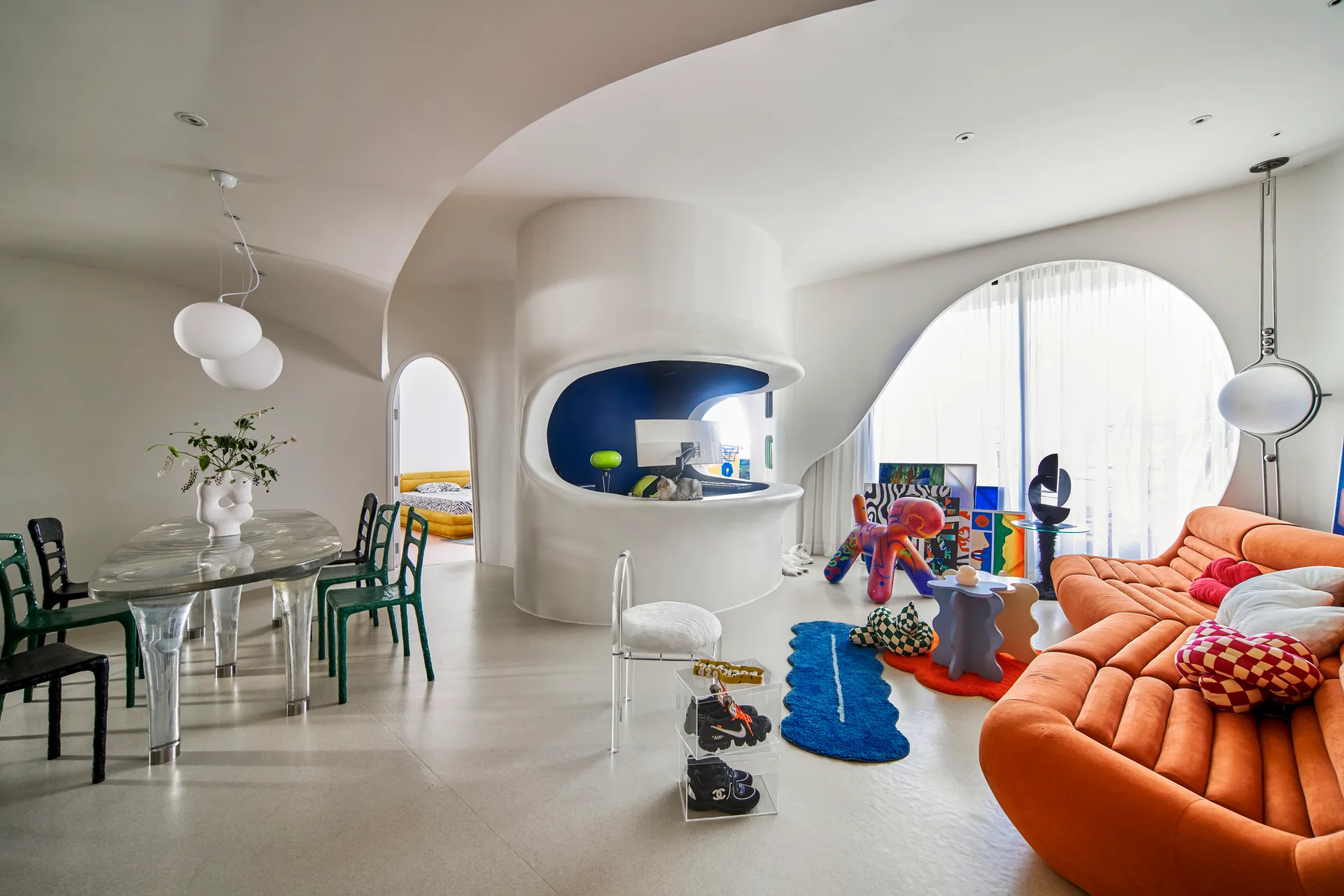 A postmodern living and dining space features curved plaster walls, a large orange channel-tufted sofa, and an oval metallic dining table.