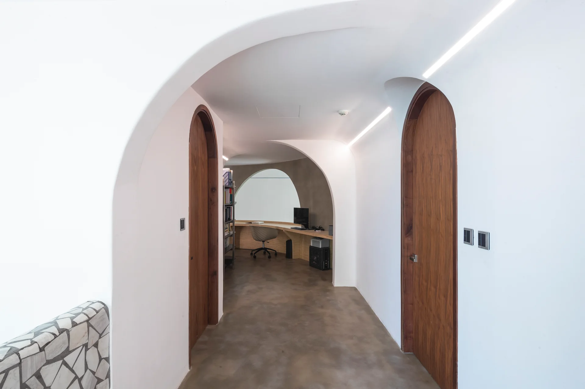 A white arched hallway with smooth plaster walls, recessed linear lights, and arched wooden doors leads to a home office with a curved desk.
