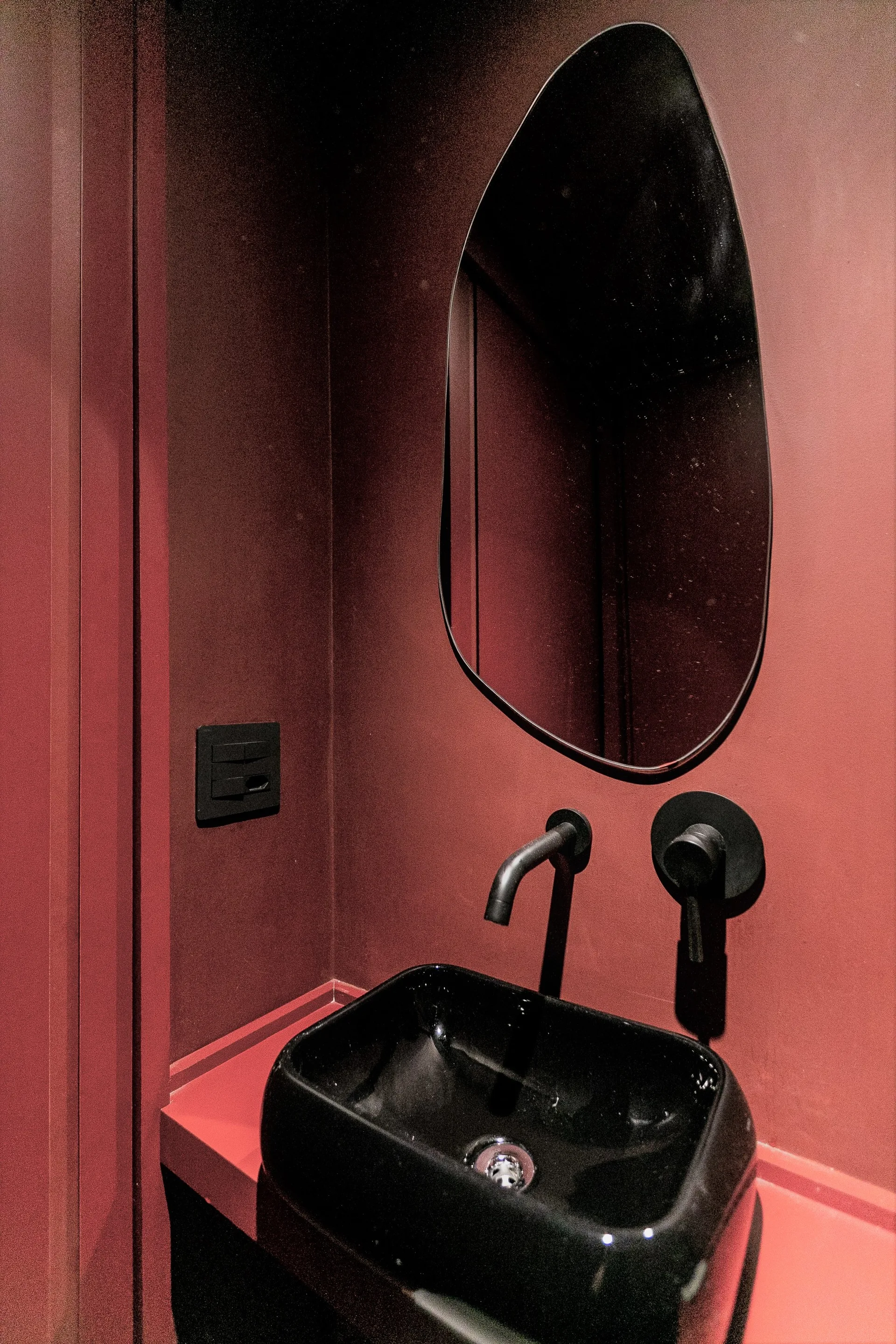 A black vessel sink and a black wall-mounted faucet are set against burgundy walls under an organic-shaped mirror in a minimalist powder room.