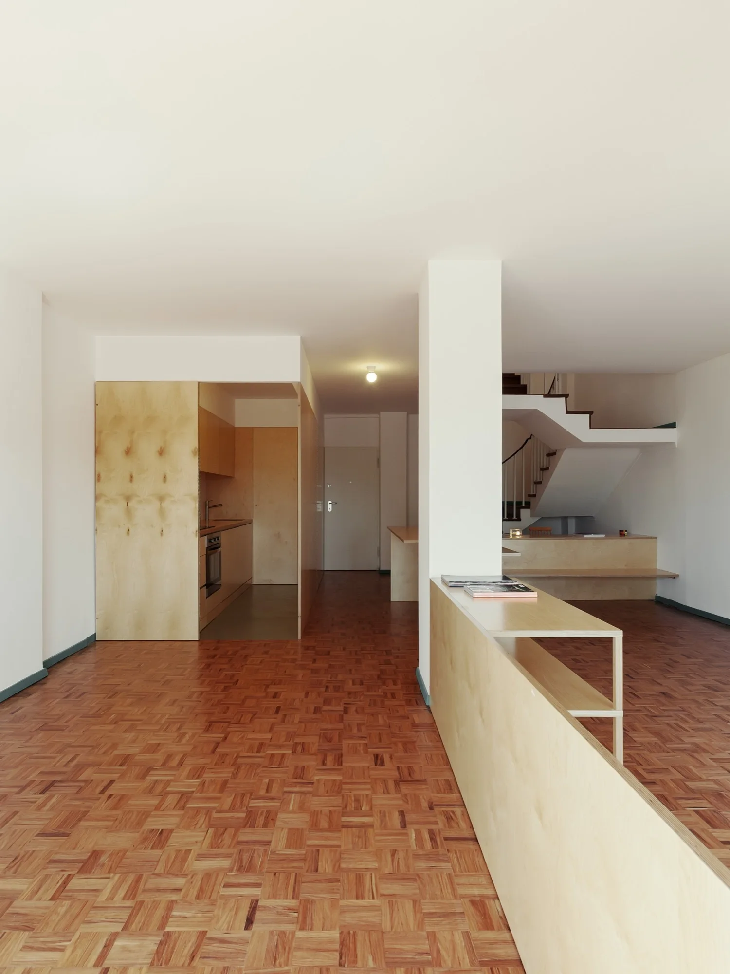 A minimalist open-plan apartment interior with a warm parquet floor, white walls, and custom light plywood structures including a kitchen and partition.