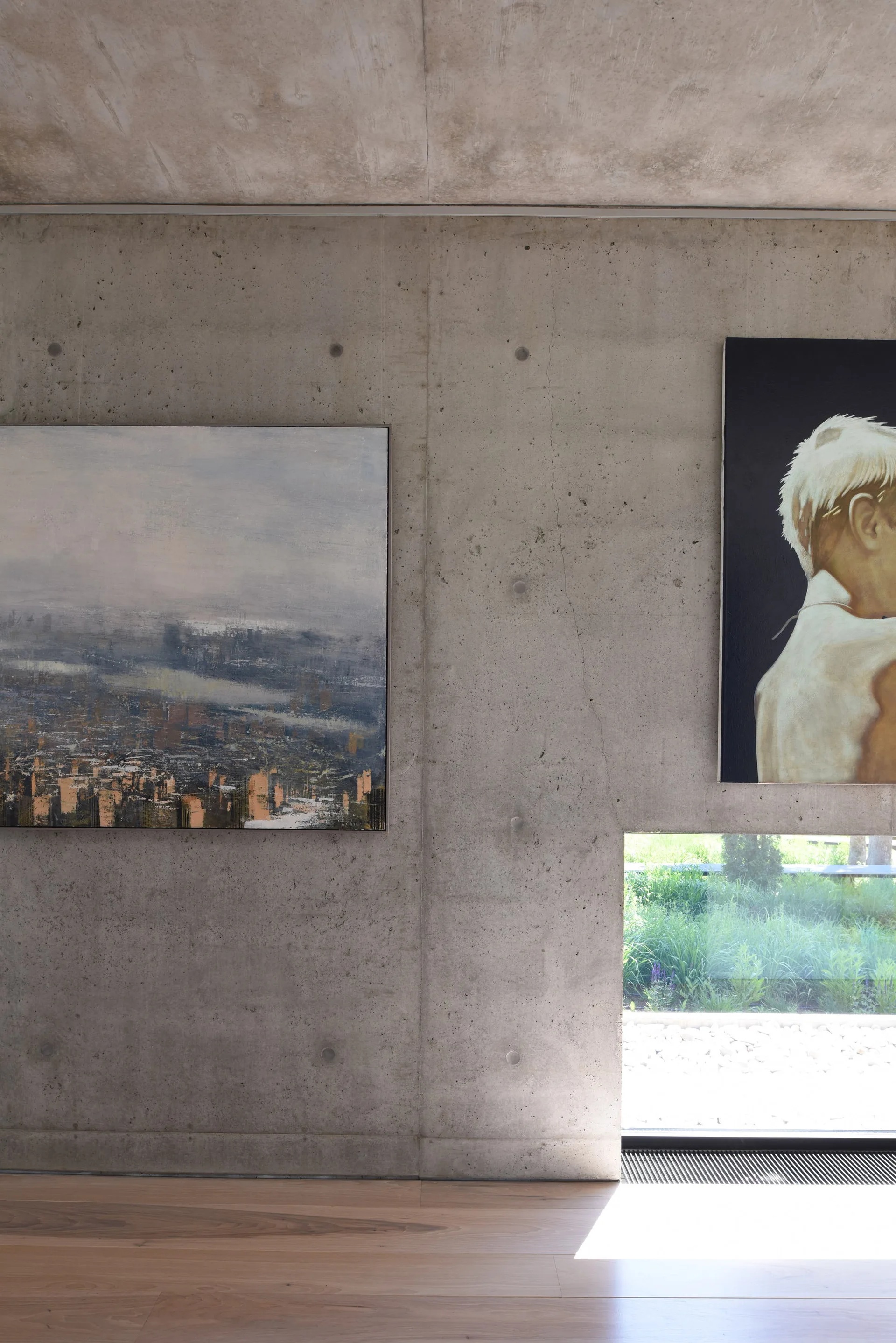 An exposed concrete wall displays two framed paintings above a light hardwood floor and a low rectangular window showing outdoor greenery.