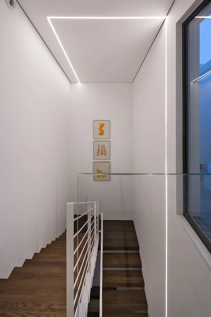 A minimalist staircase features dark wood steps, white walls, a glass railing panel, and bright linear LED light strips recessed into the ceiling.