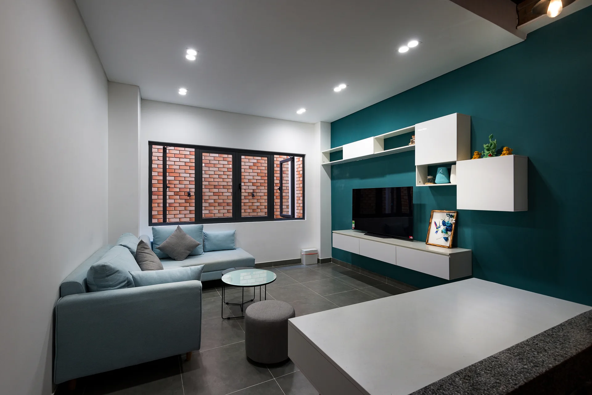 A modern living room features a light blue sectional sofa, a round glass coffee table, and a white floating console on a dark teal wall.