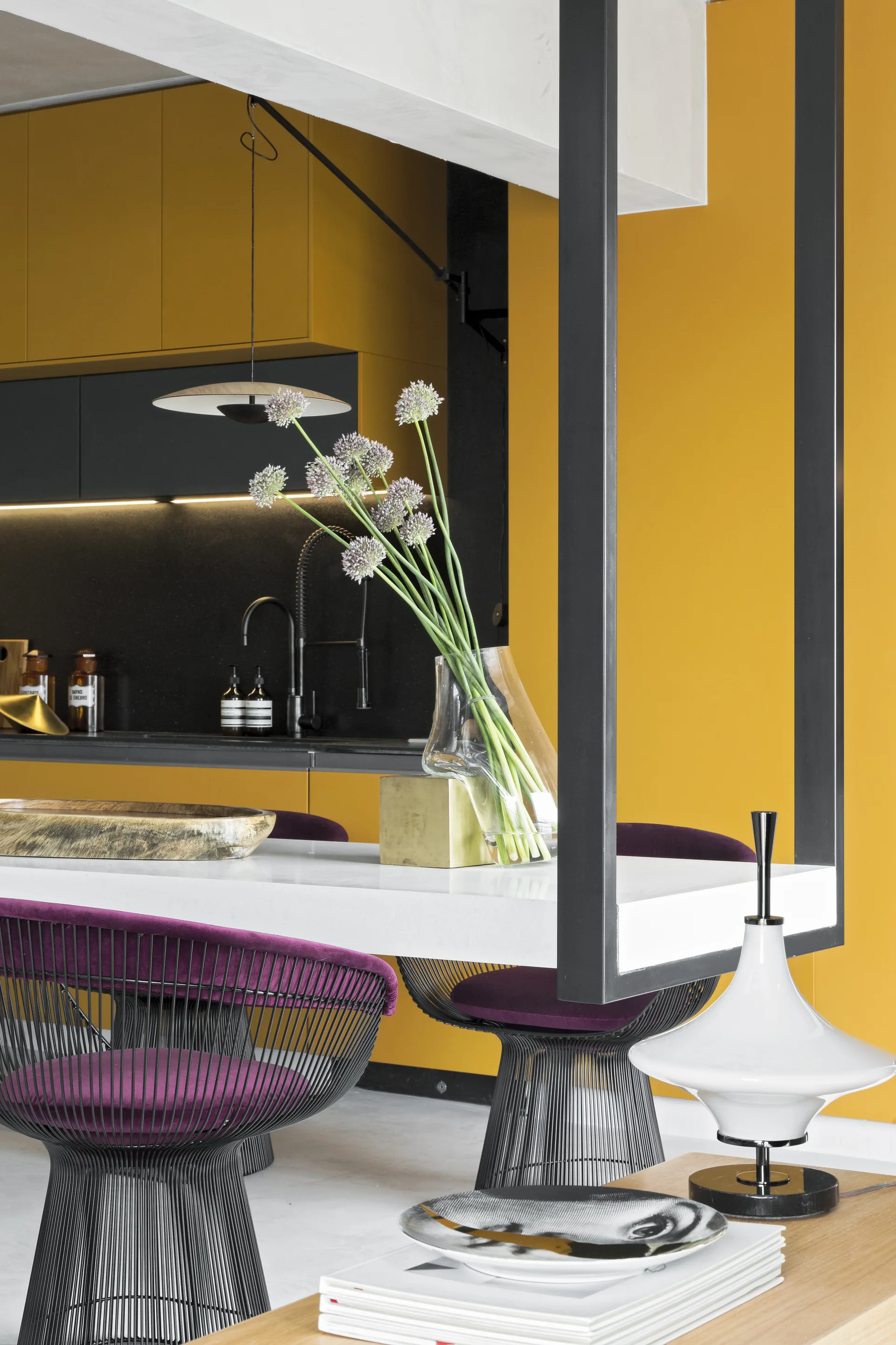 A modern kitchen space displays vibrant yellow cabinetry behind a white dining island paired with purple wire-frame chairs.