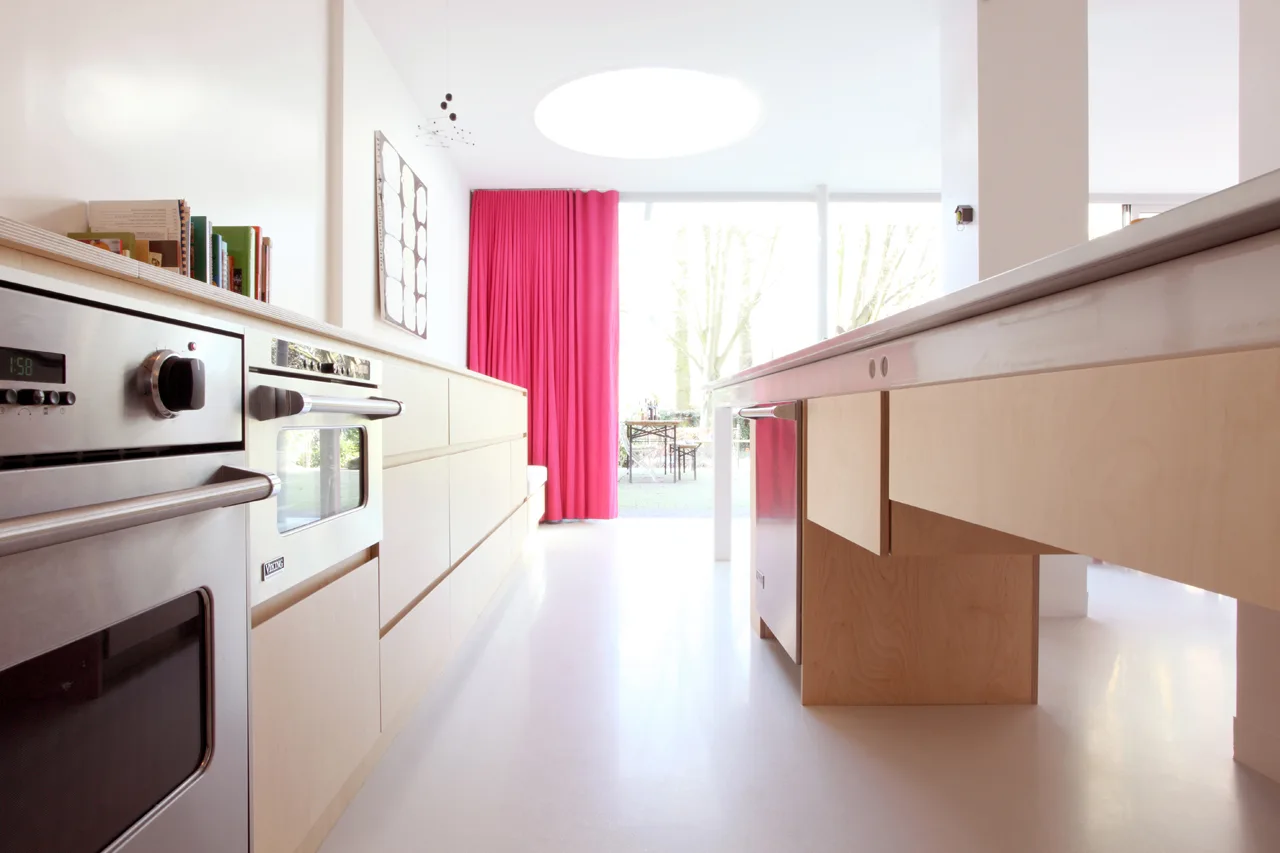 A bright kitchen features minimalist light wood cabinets, stainless steel appliances, a circular ceiling skylight, and a vibrant magenta curtain.