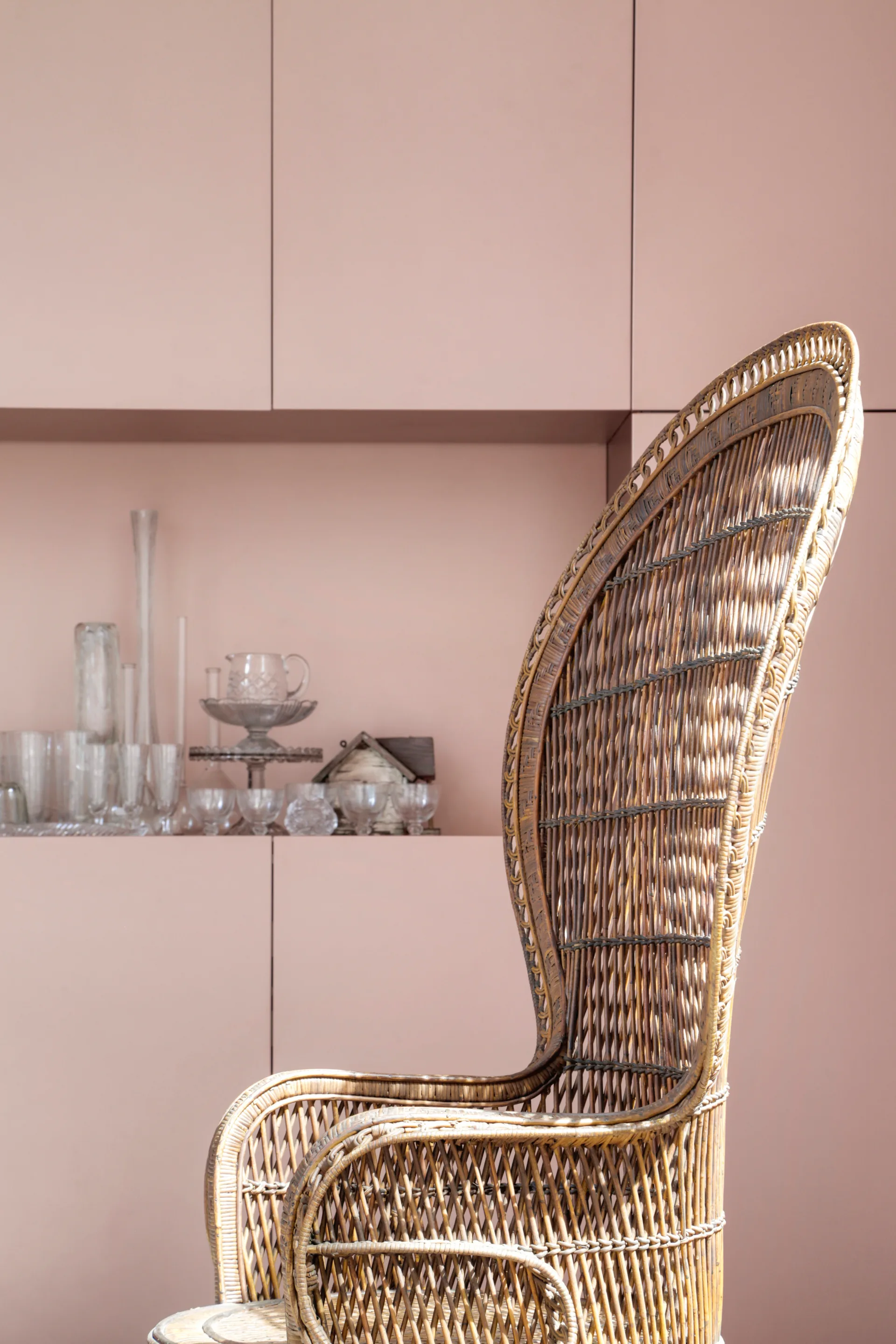A vintage woven wicker peacock chair sits in front of a matte pink built-in cabinet wall with a recessed shelf displaying clear glassware.