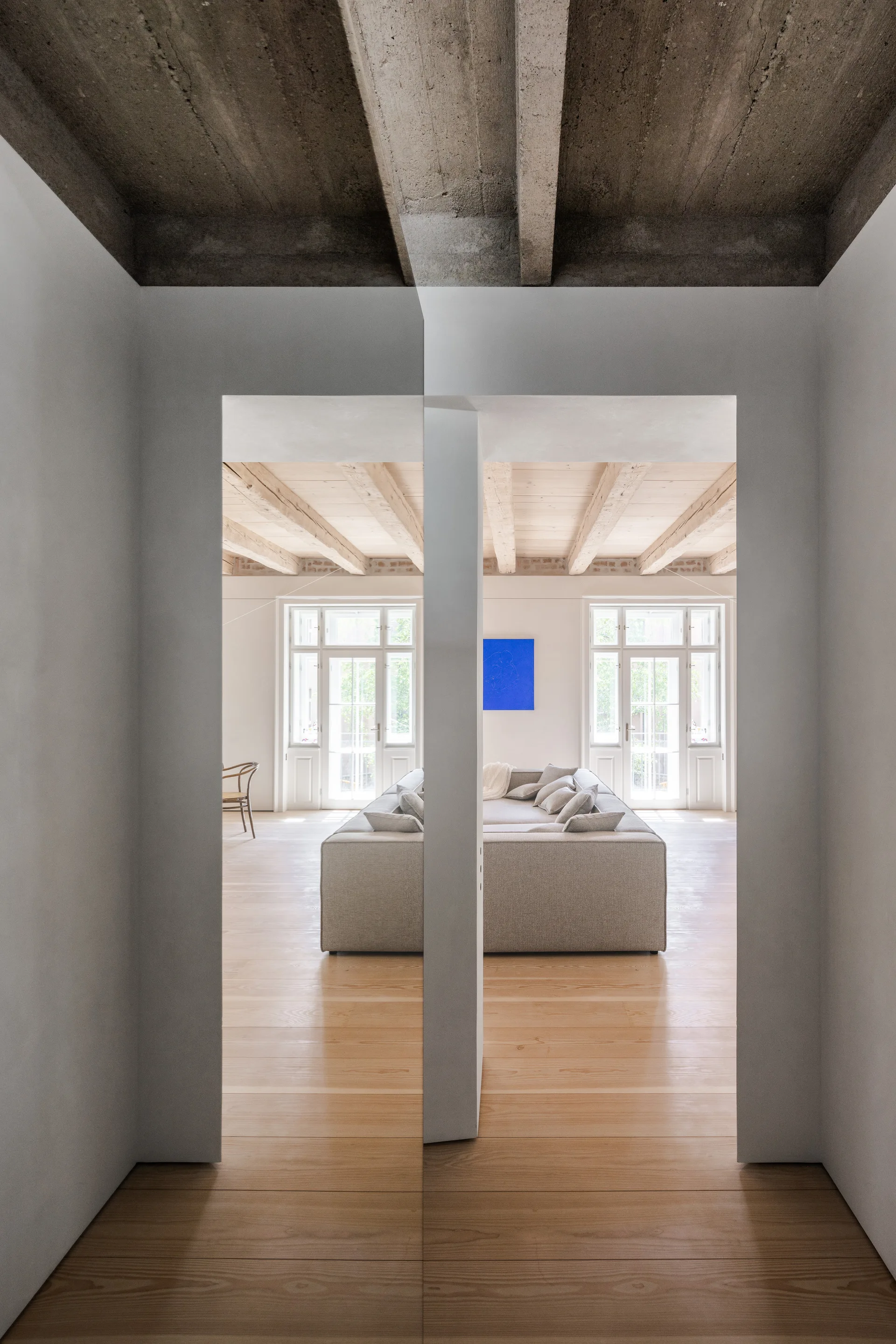 A minimalist living room with a grey sofa and exposed wooden beams, viewed through a narrow white doorway under a concrete ceiling.