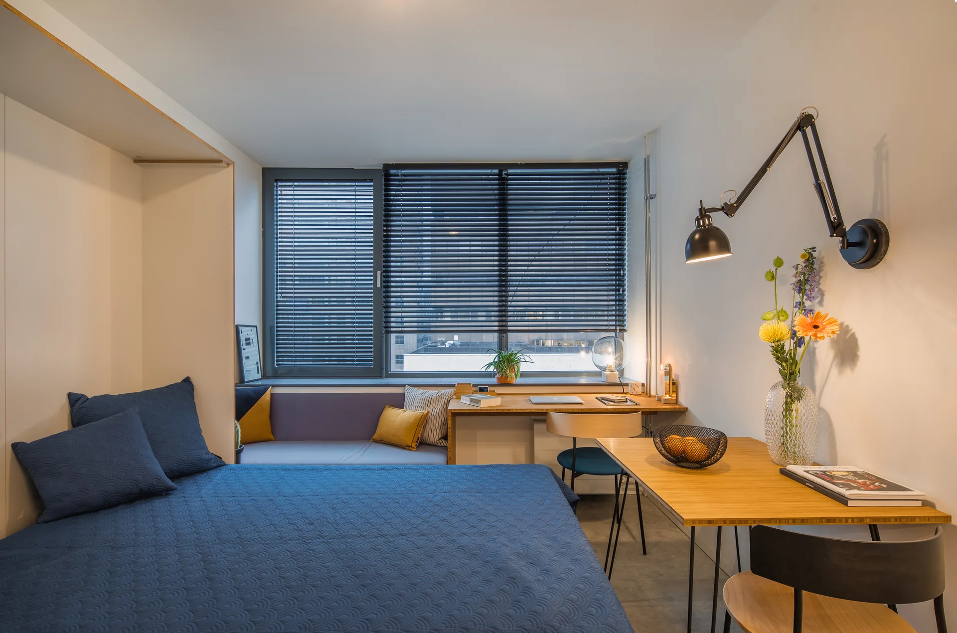 A compact bedroom features a bed with blue quilted bedding next to an integrated wooden desk and a window bench with dark venetian blinds.