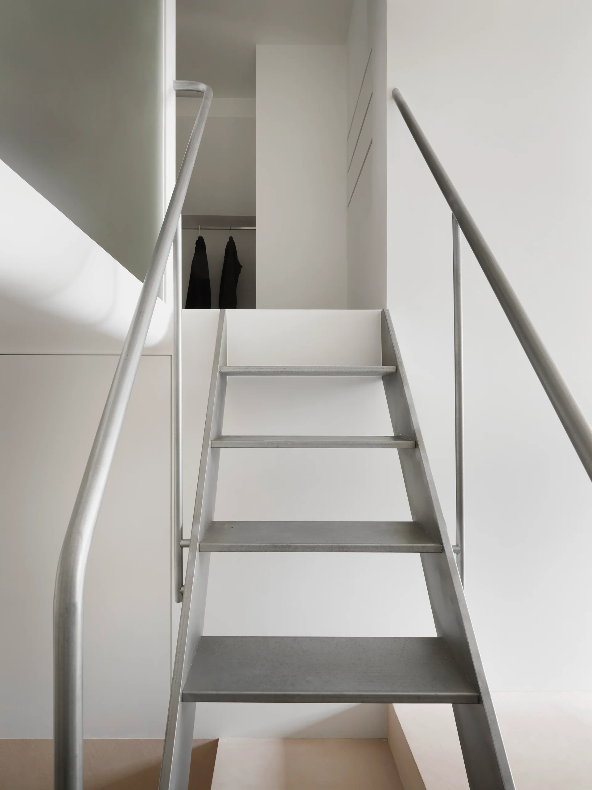 A minimalist white interior featuring a matte metal staircase with round handrails leading up to an open doorway.
