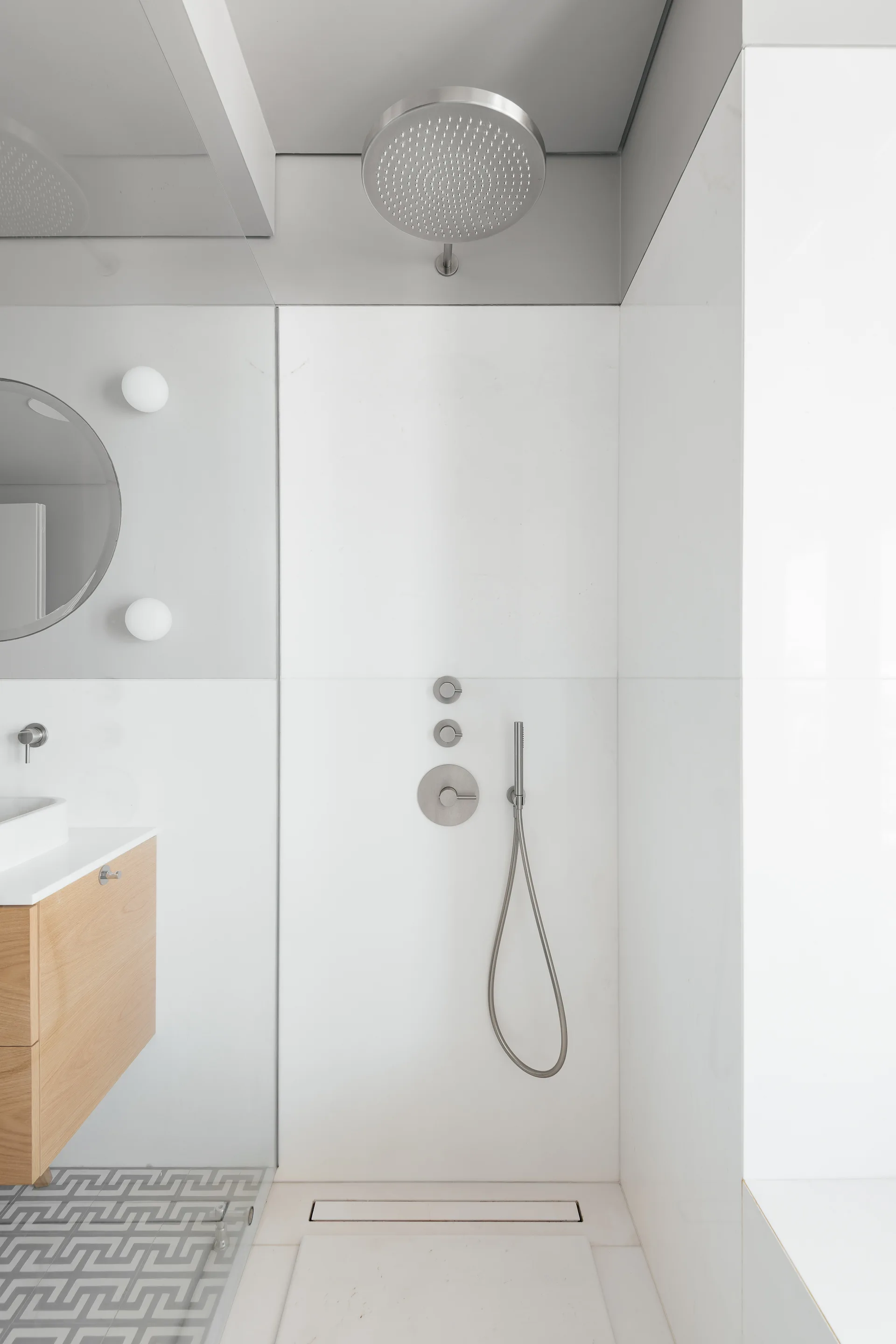 A minimalist bathroom features a white walk-in shower with brushed metal hardware next to a floating wooden vanity and geometric floor tiles.