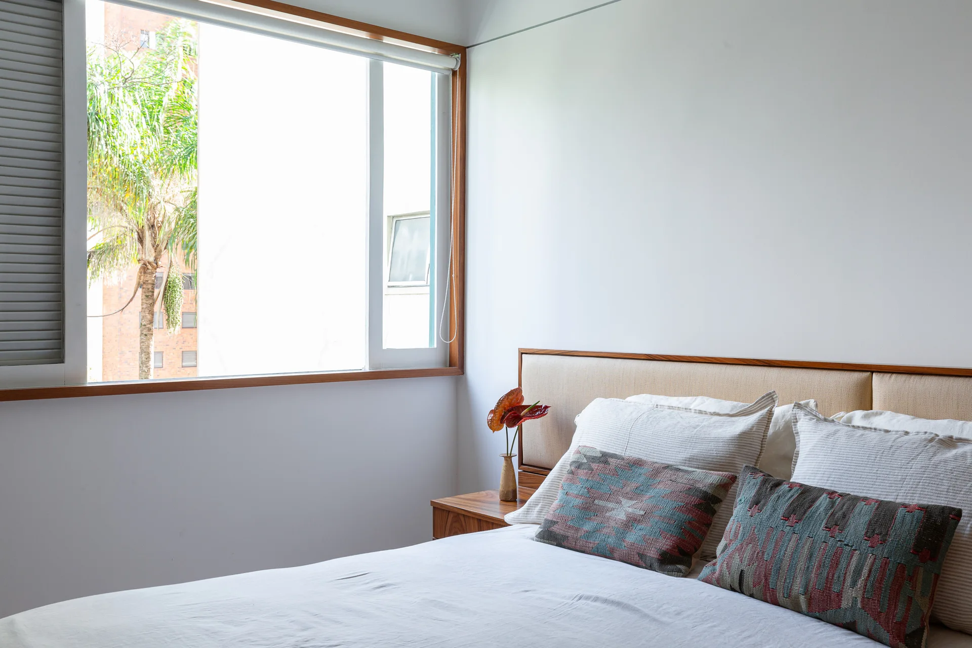 A minimalist bedroom features a bed with patterned accent pillows and a wooden headboard next to a window framed in warm wood trim.