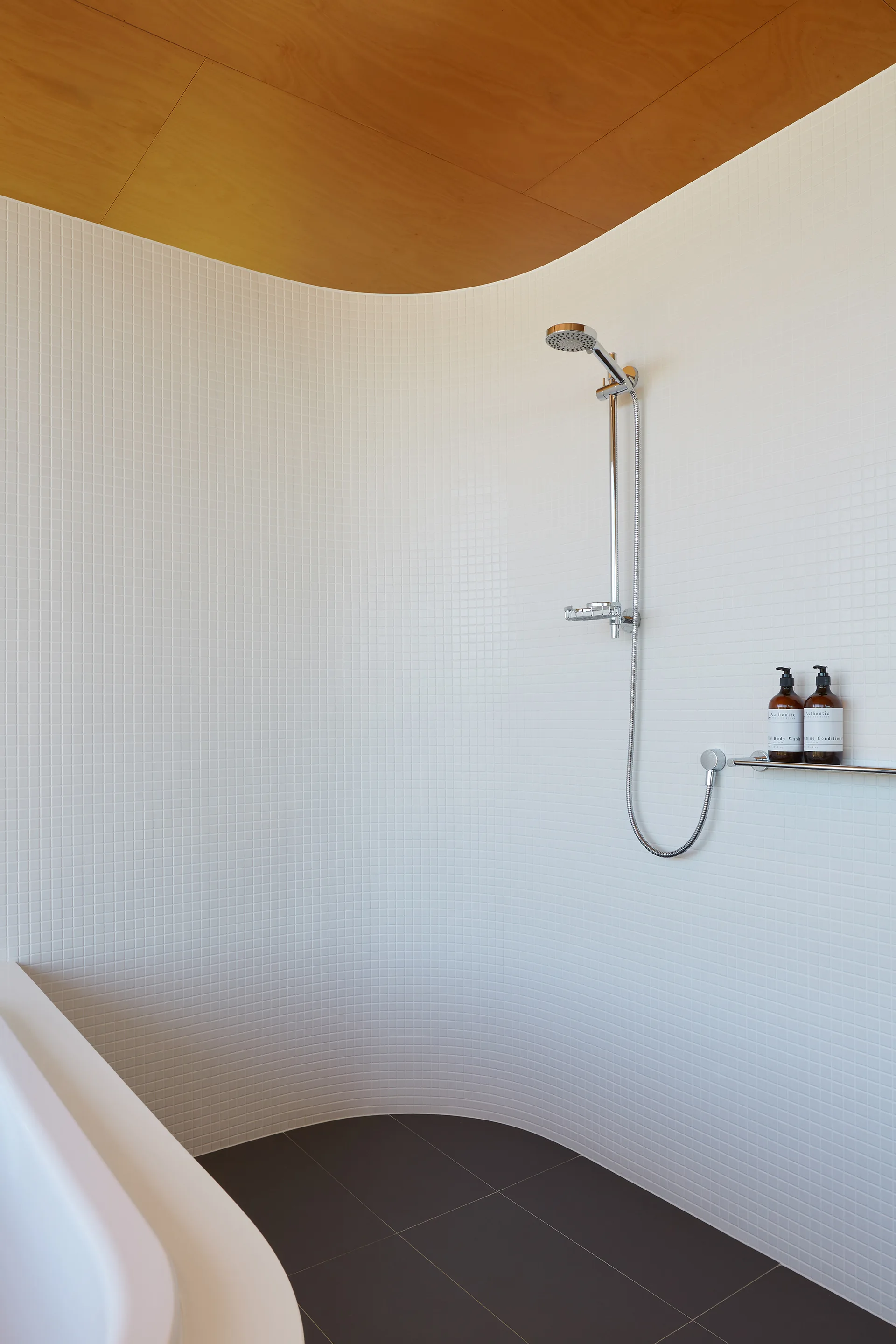 A modern minimalist shower features a curved wall of small white tiles, a natural wood ceiling, and chrome fixtures.