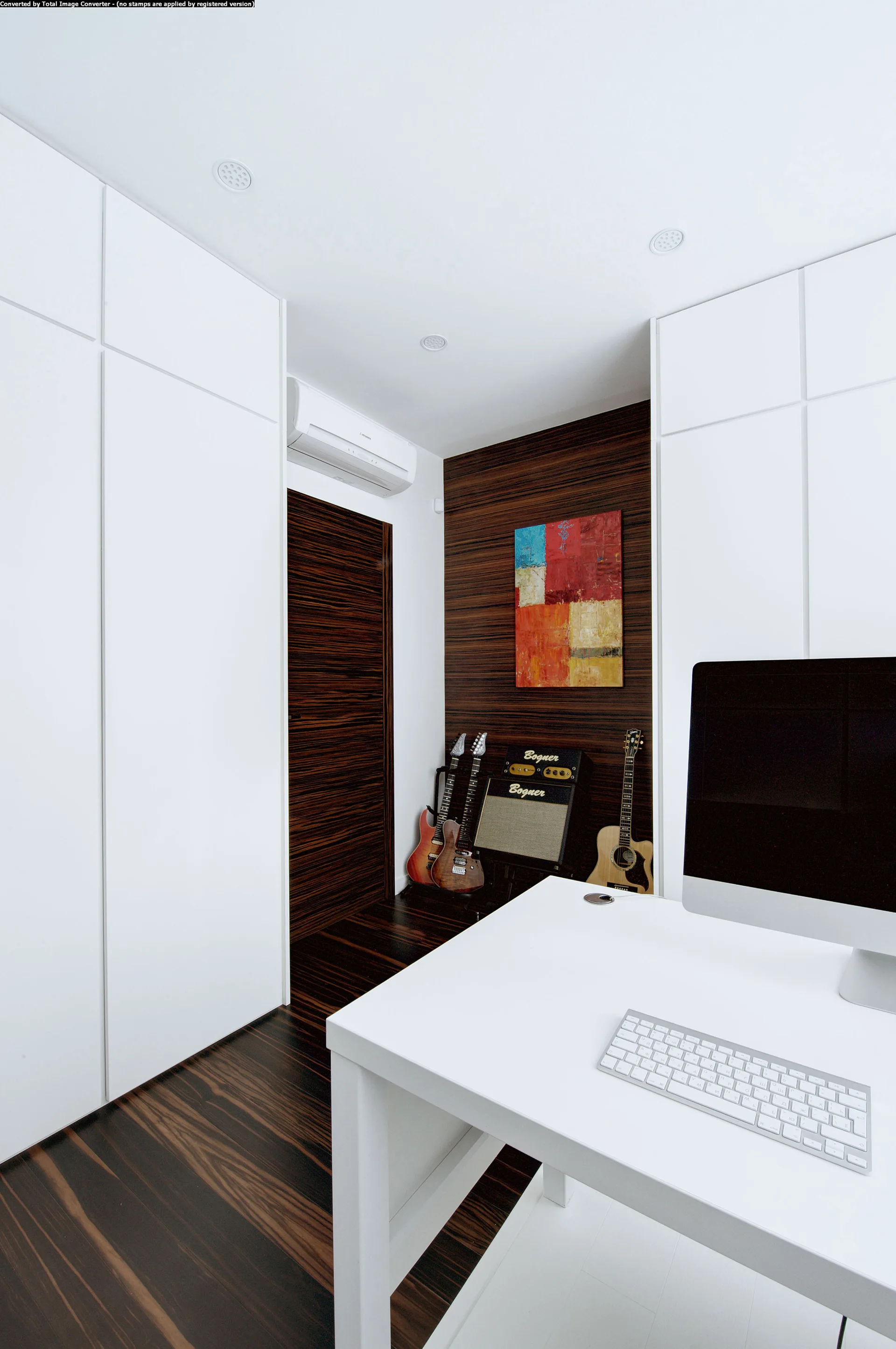 A minimalist study featuring a white desk, large built-in white cabinets, and a dark wood accent wall with displayed guitars.
