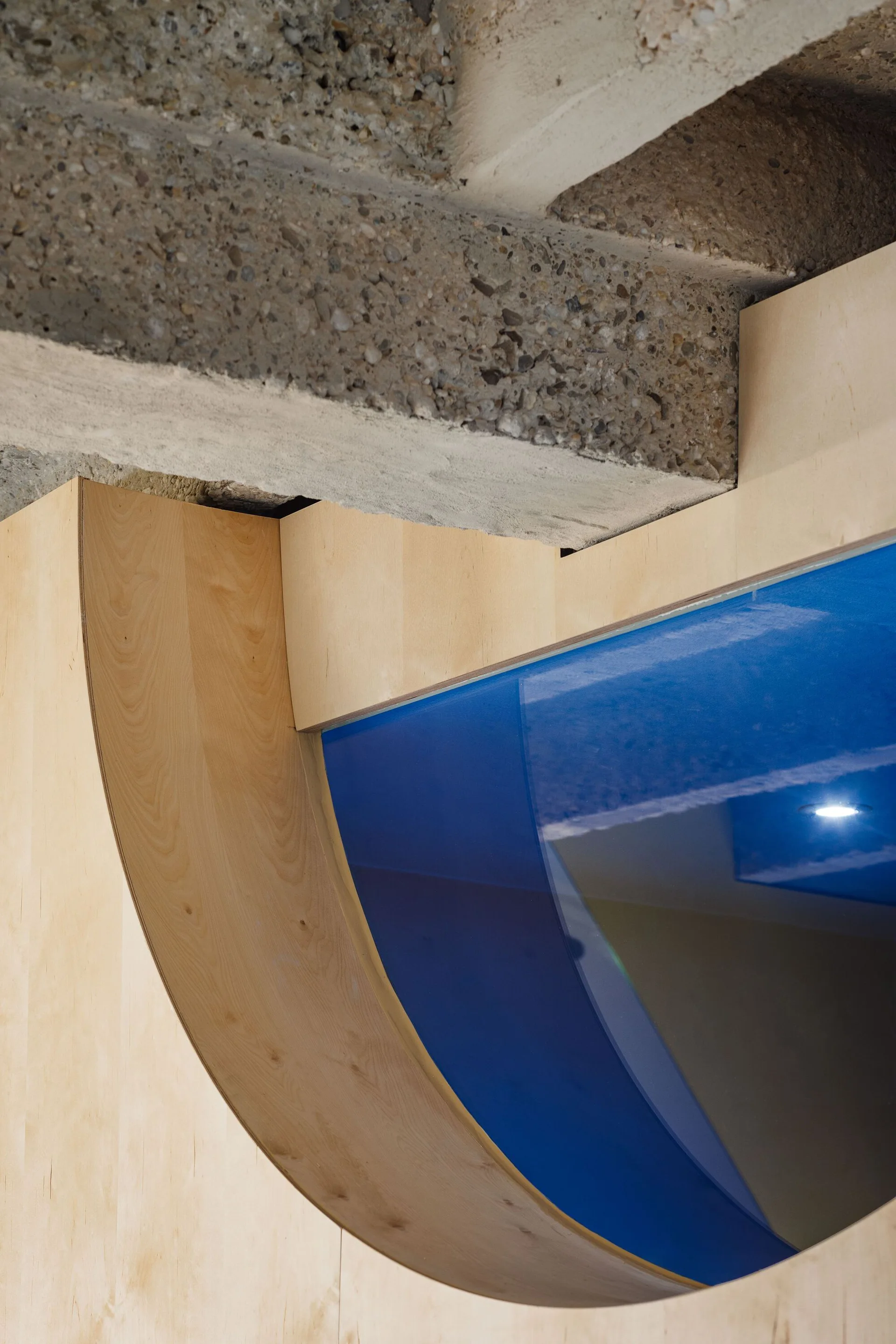 An architectural close-up of a curved light wood plywood wall meeting a textured grey concrete beam next to a reflective blue panel.