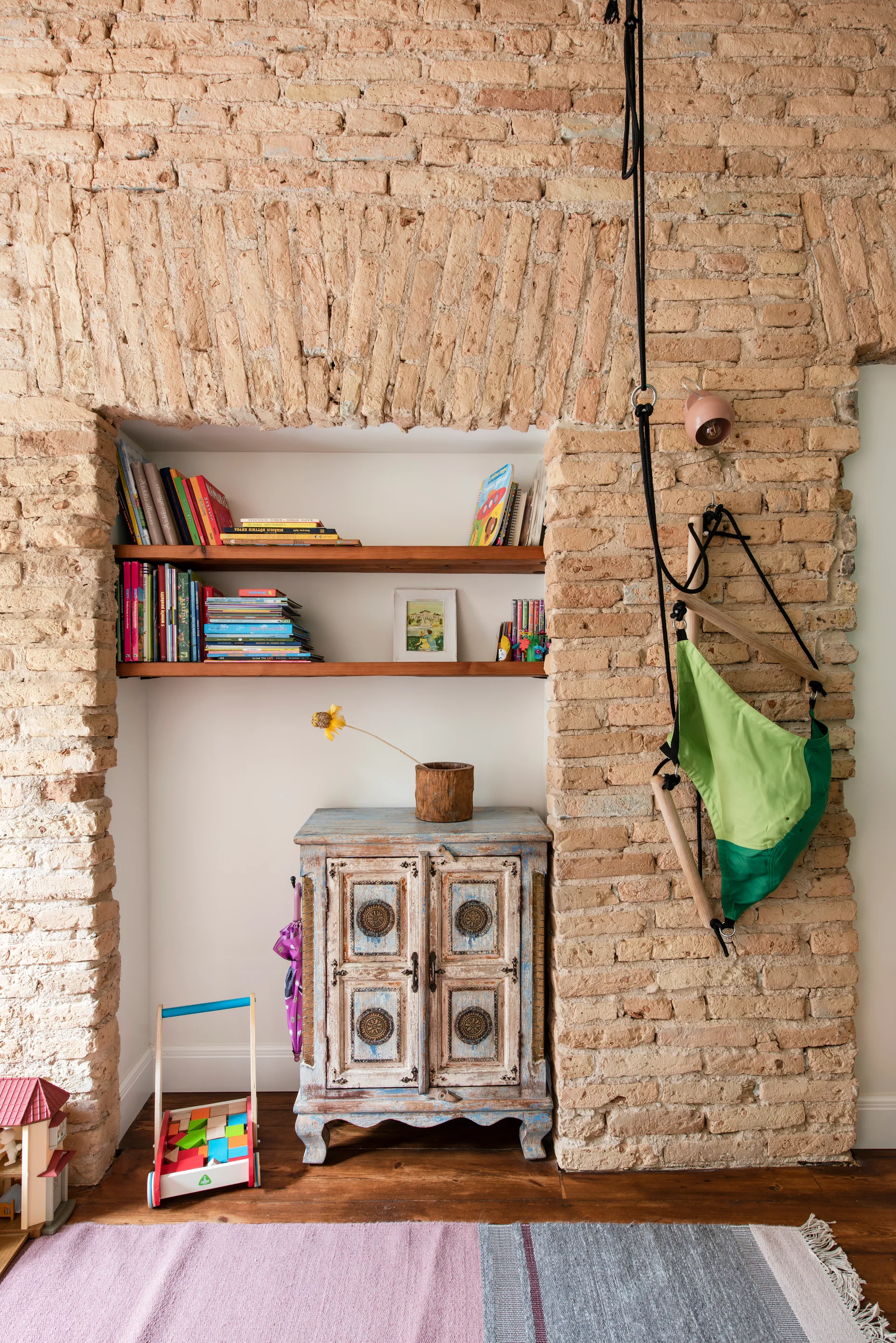A playroom alcove framed by an exposed brick archway features recessed wooden bookshelves, a distressed vintage cabinet, and a wooden floor.