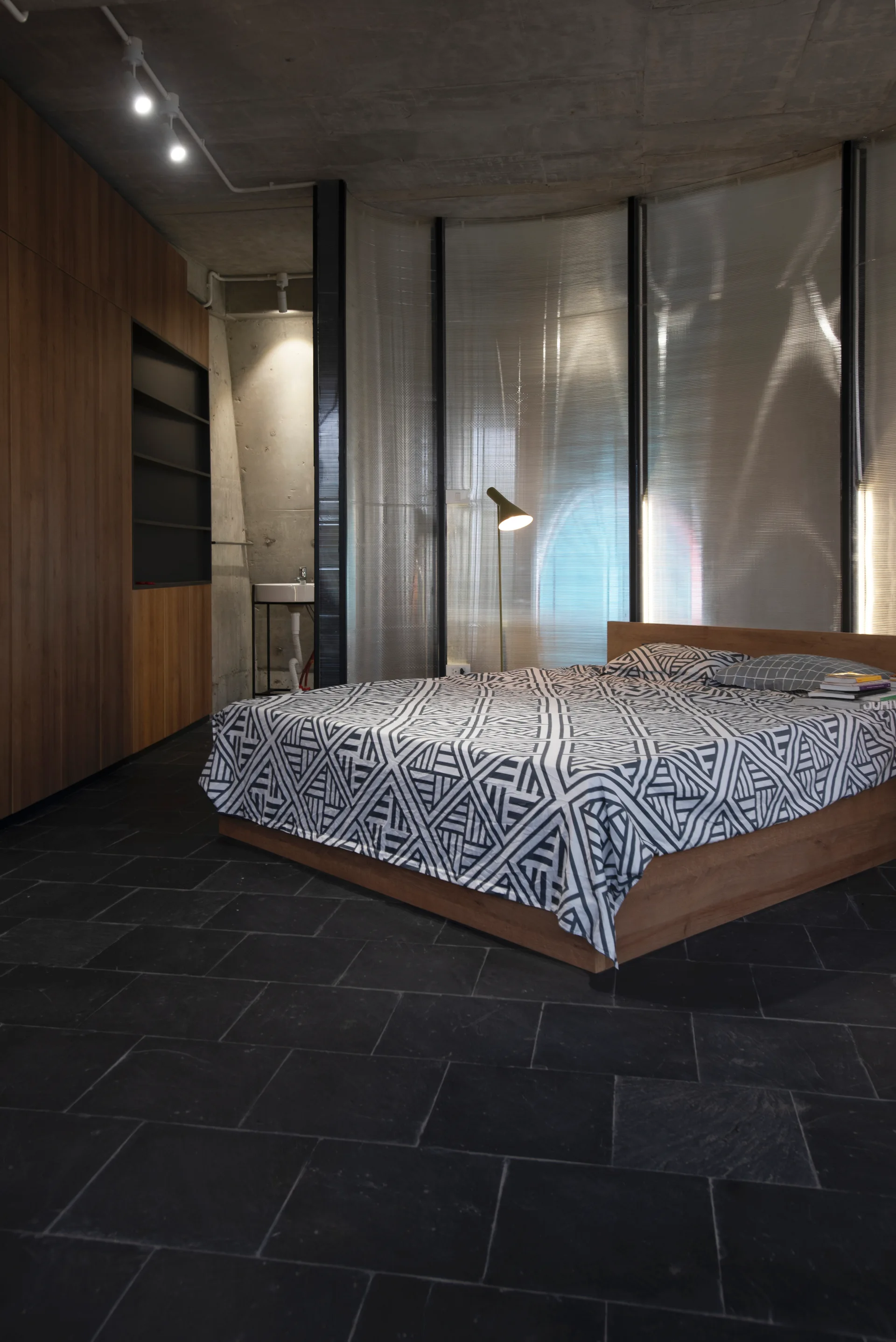A modern bedroom features a wooden bed with geometric bedding, dark slate tile flooring, raw concrete ceiling, and a curved translucent partition screen.