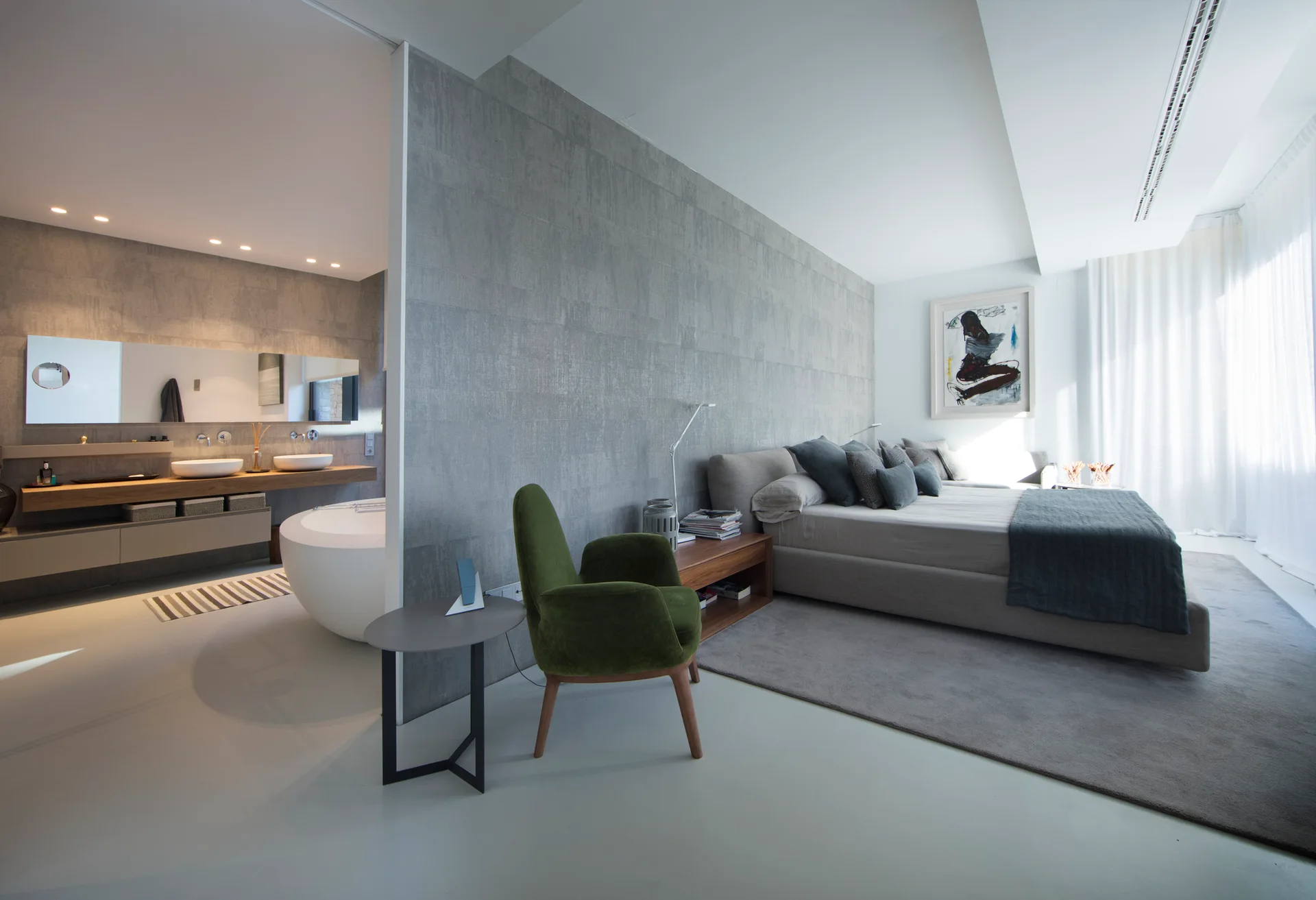 A modern primary bedroom suite featuring a grey textured partition wall that separates the upholstered bed from an open-concept bathroom.