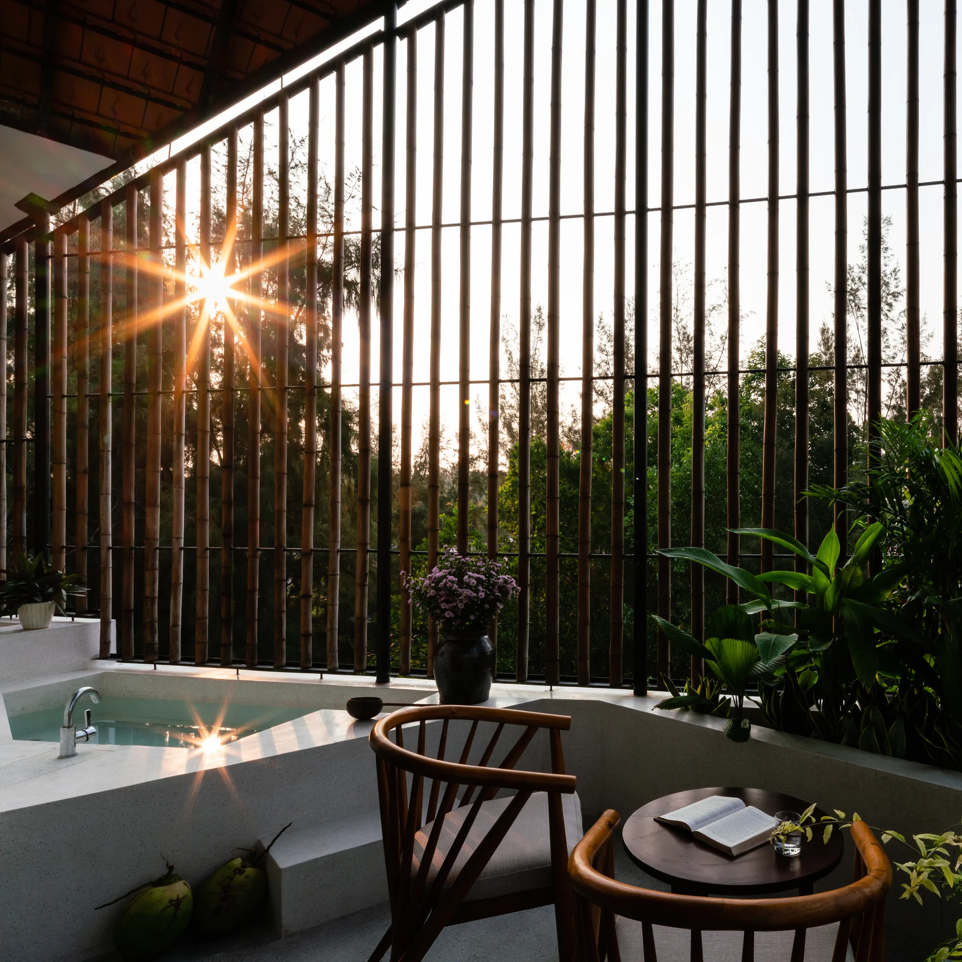 A semi-outdoor patio features a white built-in soaking tub, a vertical bamboo screen wall, and a wooden seating area under dappled sunlight.