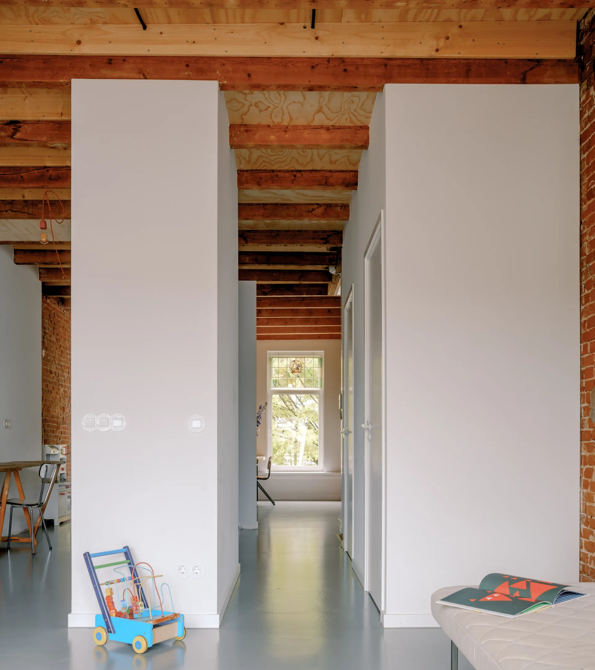 An industrial hallway featuring exposed wooden ceiling beams, white structural partitions, textured red brick walls, and a smooth gray floor.