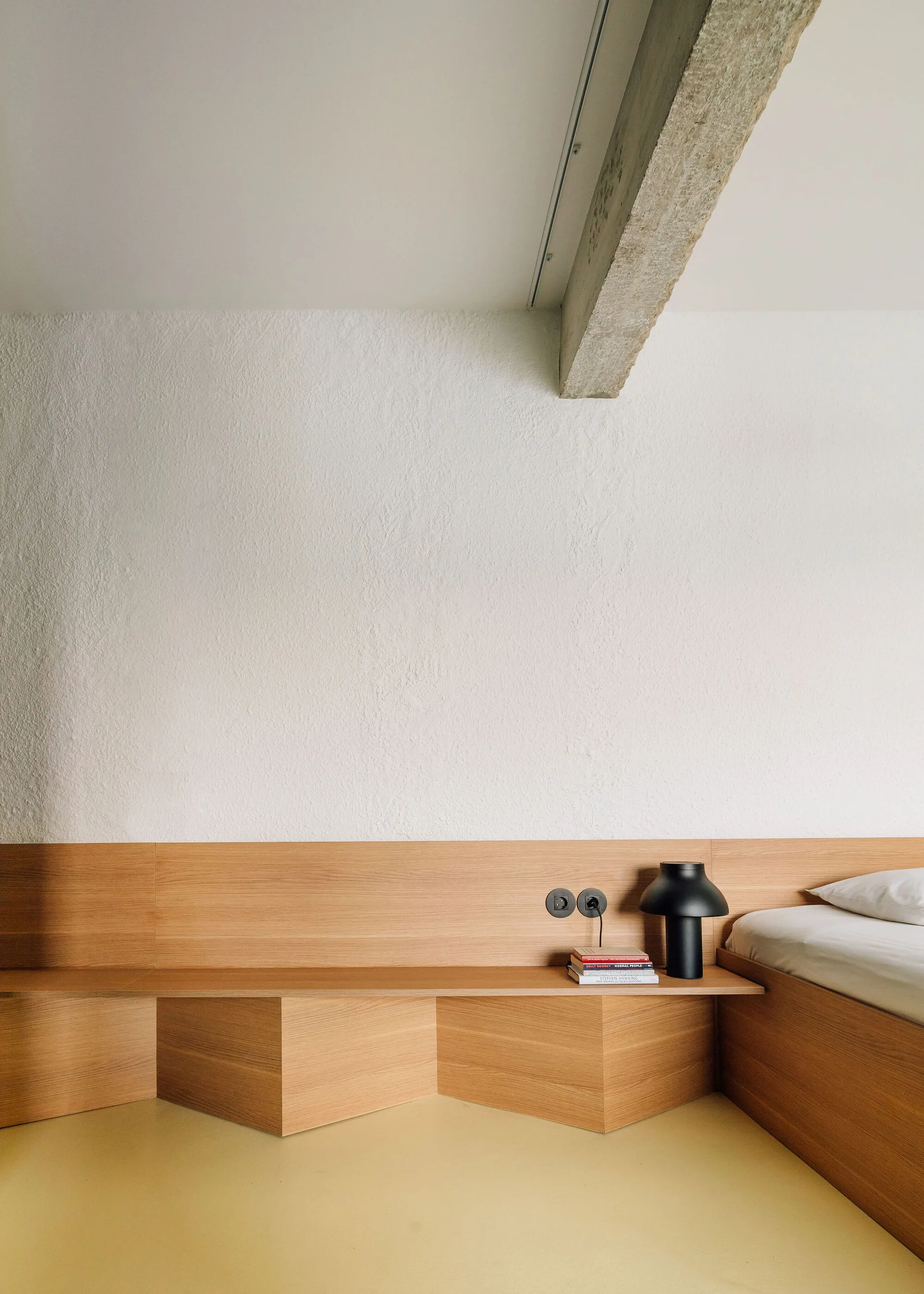 A minimalist bedroom features a custom wooden headboard extending into a low shelf against a textured plaster wall with an exposed concrete ceiling beam.