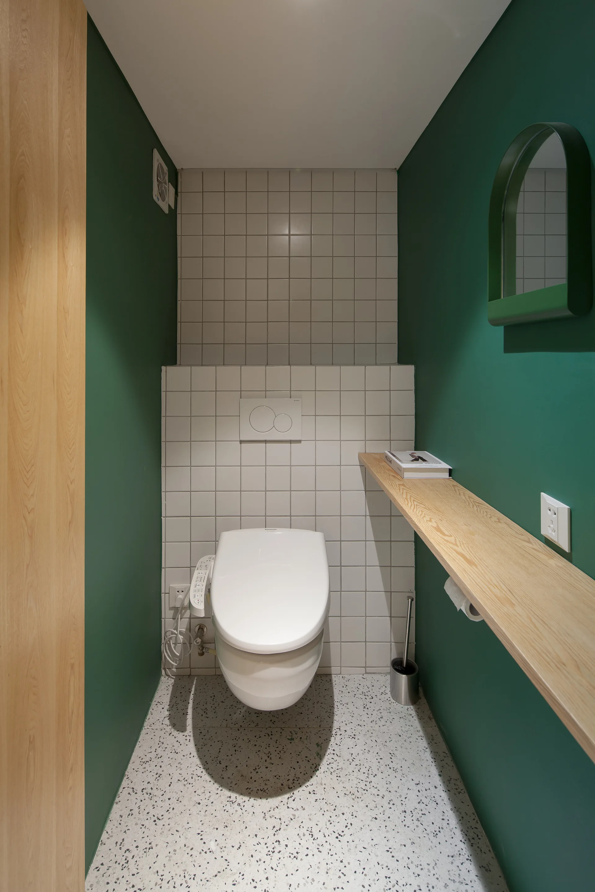 A small powder room features a white wall-hung toilet against grid tiling, green walls, a light wood shelf, and terrazzo flooring.