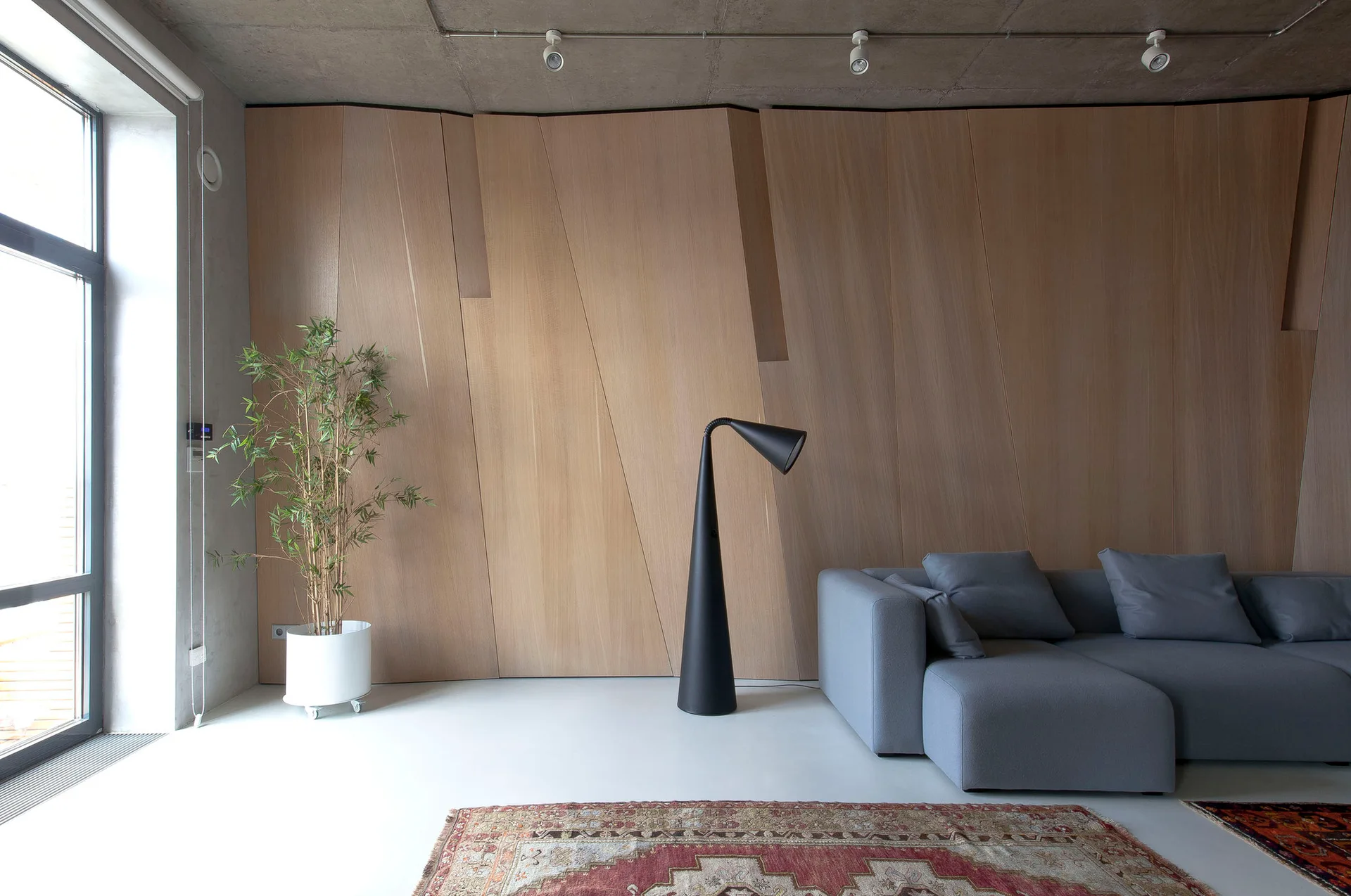 A grey modular sofa sits in front of a geometric wood-paneled wall with a minimalist black floor lamp and concrete ceiling.