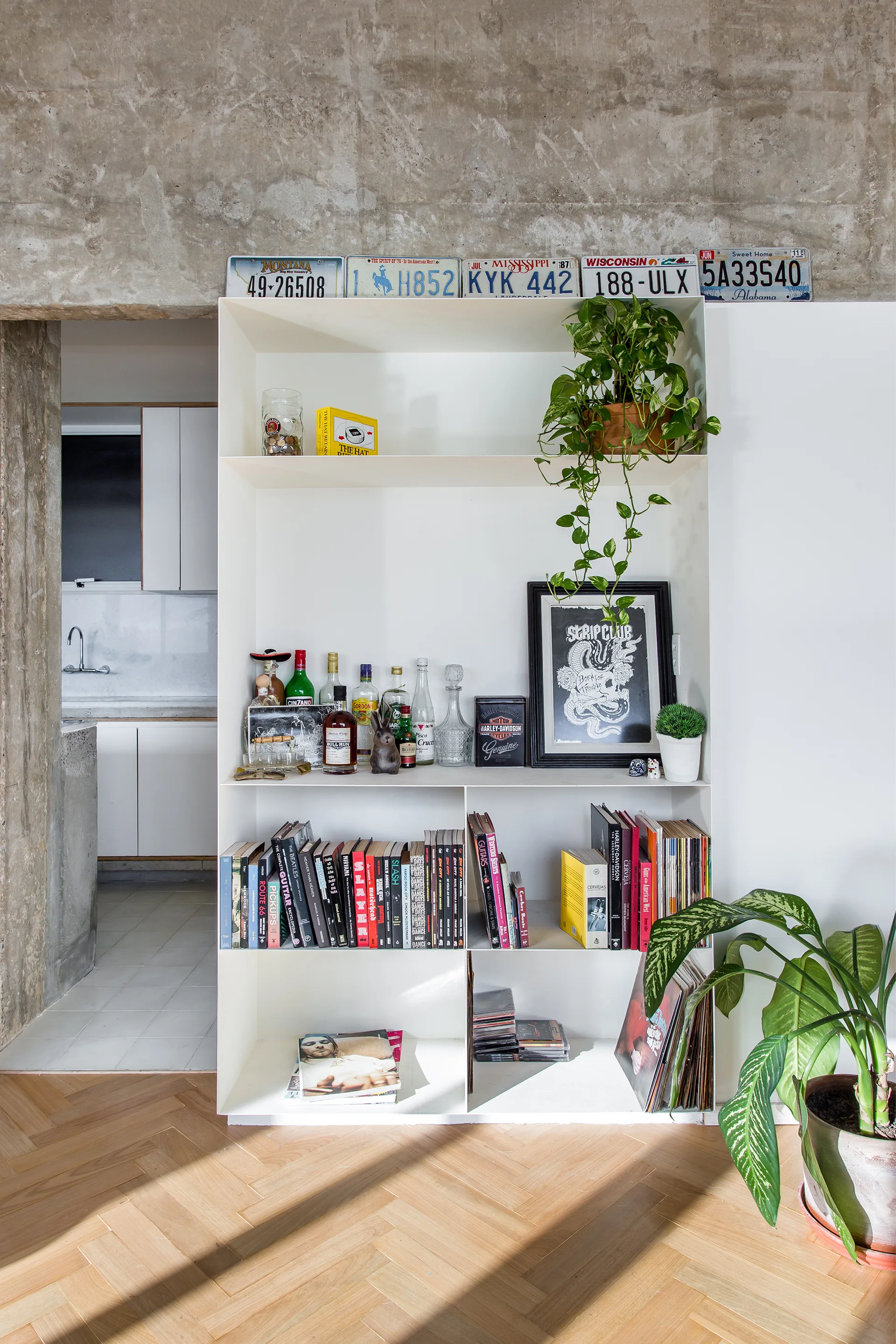 A white metal shelving unit with books, plants, and bottles sits beneath a concrete ceiling beam on herringbone wood floors.