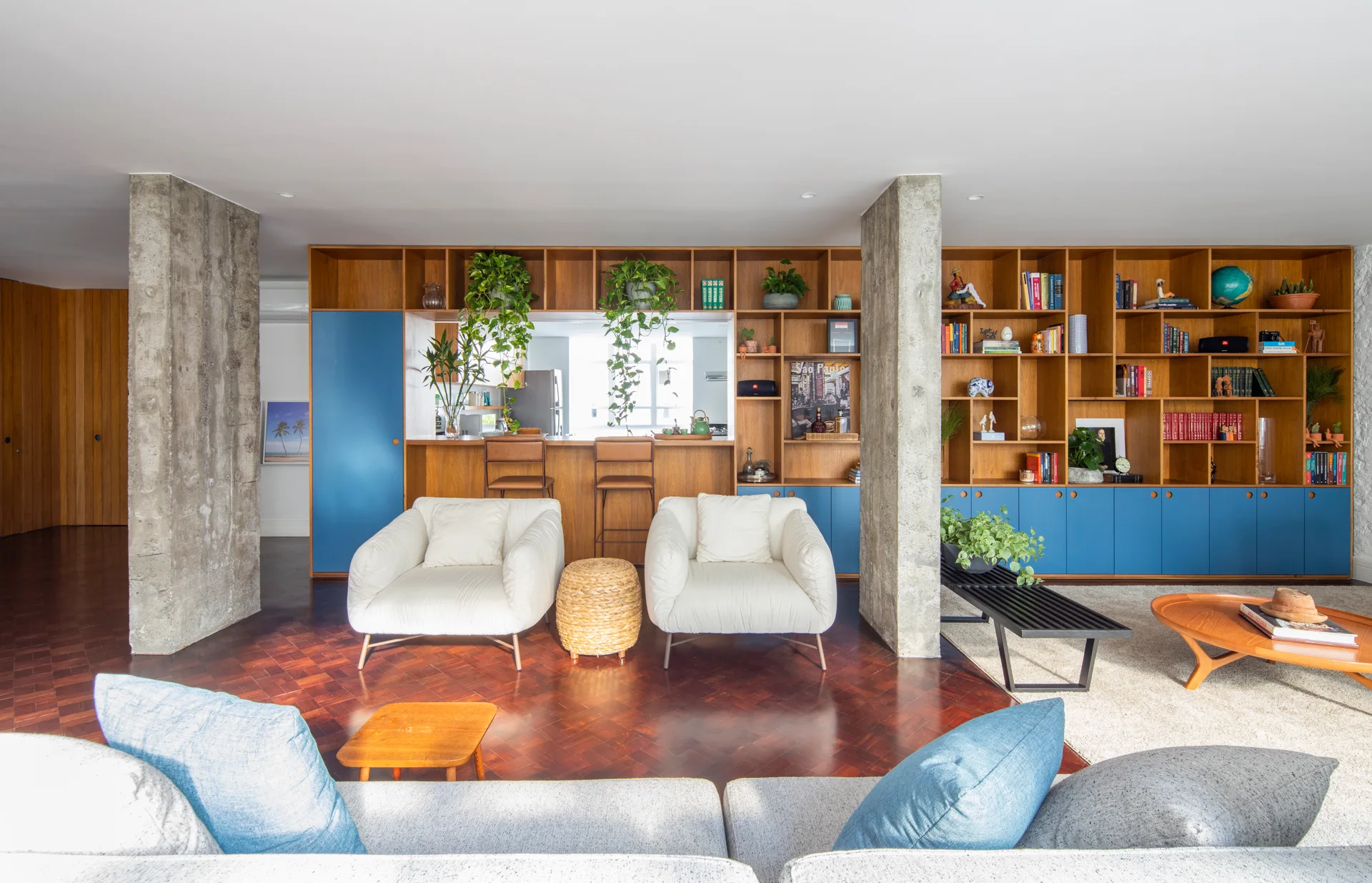 A living room features two white lounge chairs facing a custom wood shelving unit with blue cabinets, set between two exposed concrete pillars.
